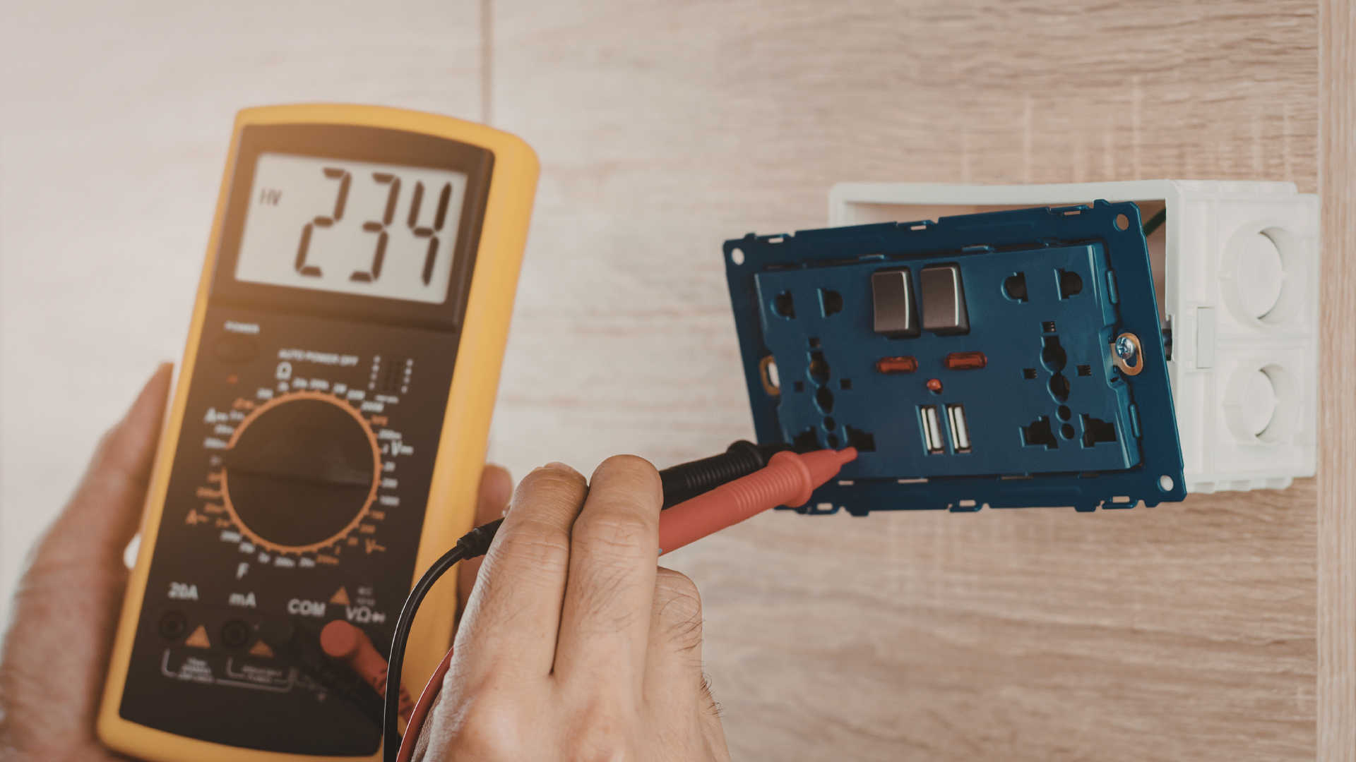 Hand holding a multimeter probes an open blue electrical wall outlet box showing 2.34 volts.