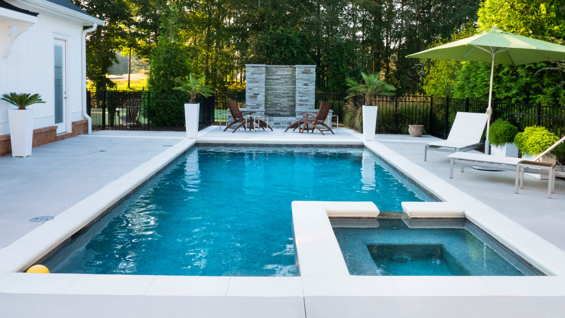 Modern backyard pool with spa, white loungers, and patio beside a house, framed by trees