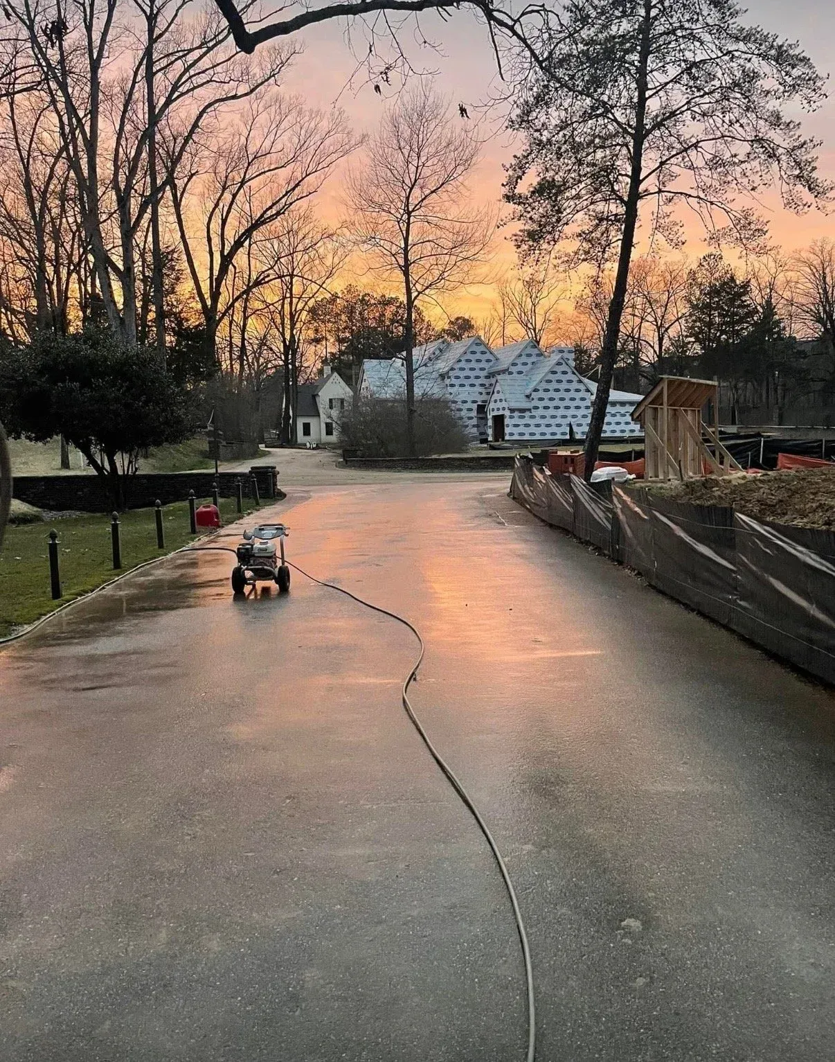 A small piece of landscaping equipment sits on a wet, paved driveway at sunrise, with a construction site in the distance.