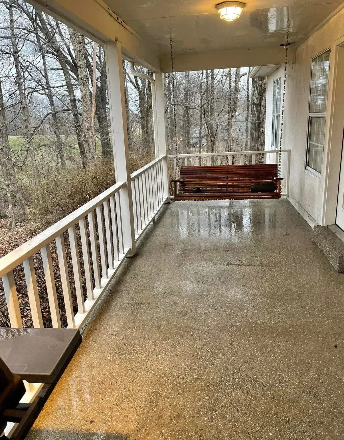 A view of a wet, covered porch with white railings, a brown hanging bench, and a wooded background on a cloudy day.