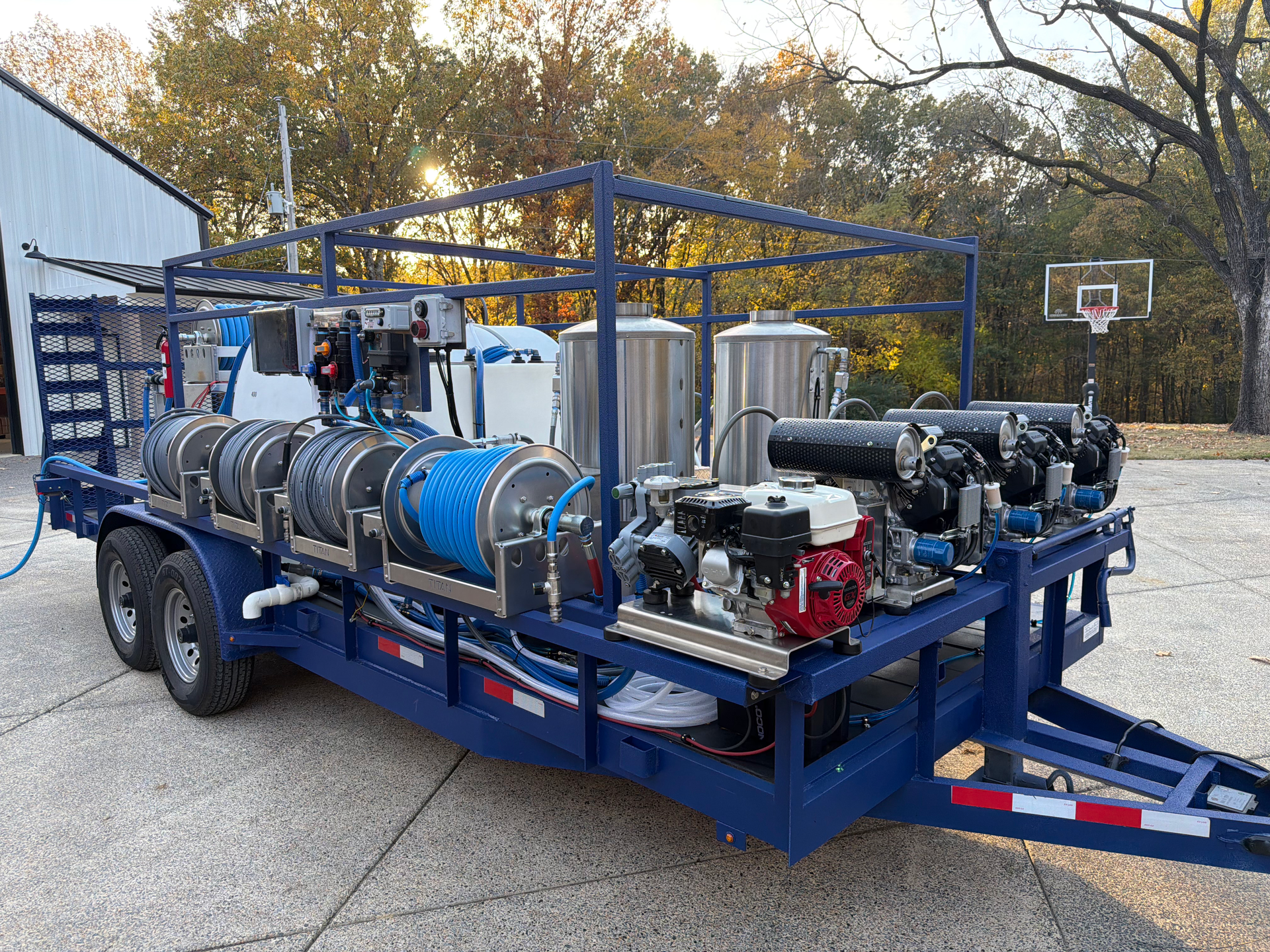 A blue utility trailer holds two silver tanks, multiple hose reels, and several engine-driven pump units outdoors.