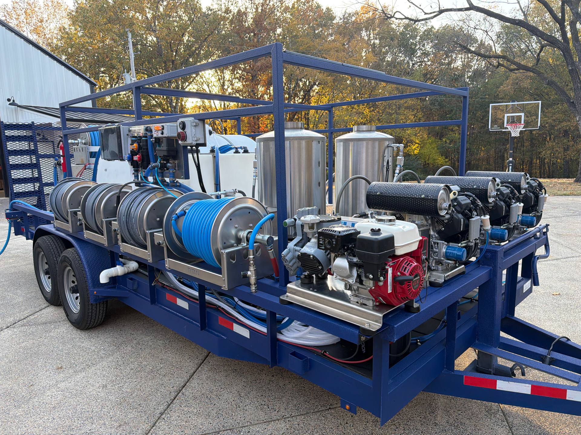 A blue utility trailer with hoses, two stainless steel tanks, and small engine-powered pressure washing equipment.
