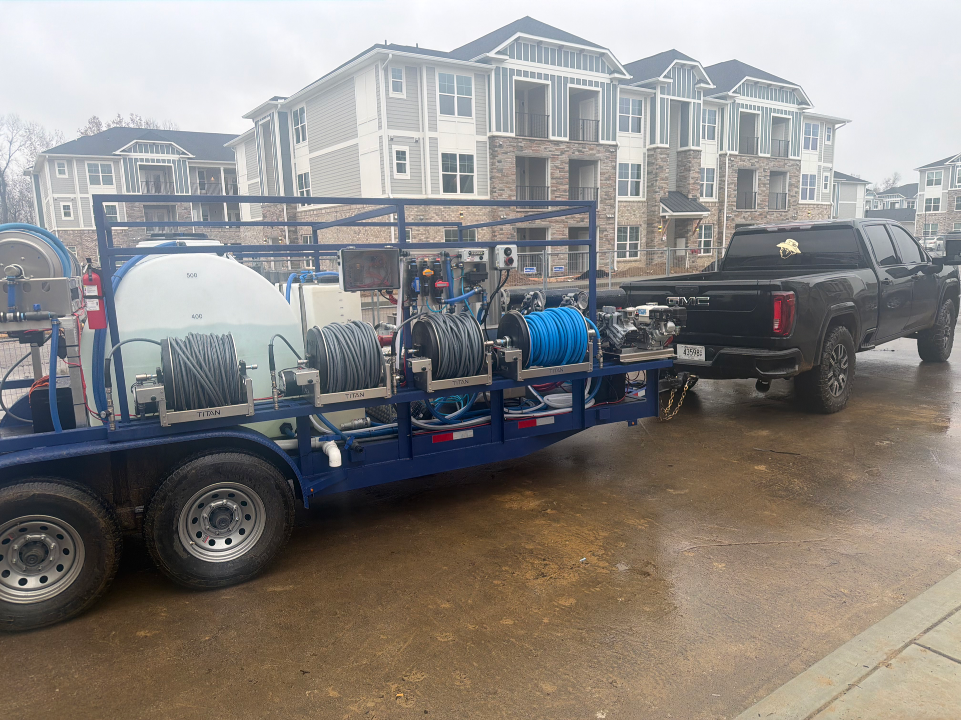 A blue trailer carrying water tanks and hose reels is hitched to a black pickup truck in front of an apartment building.