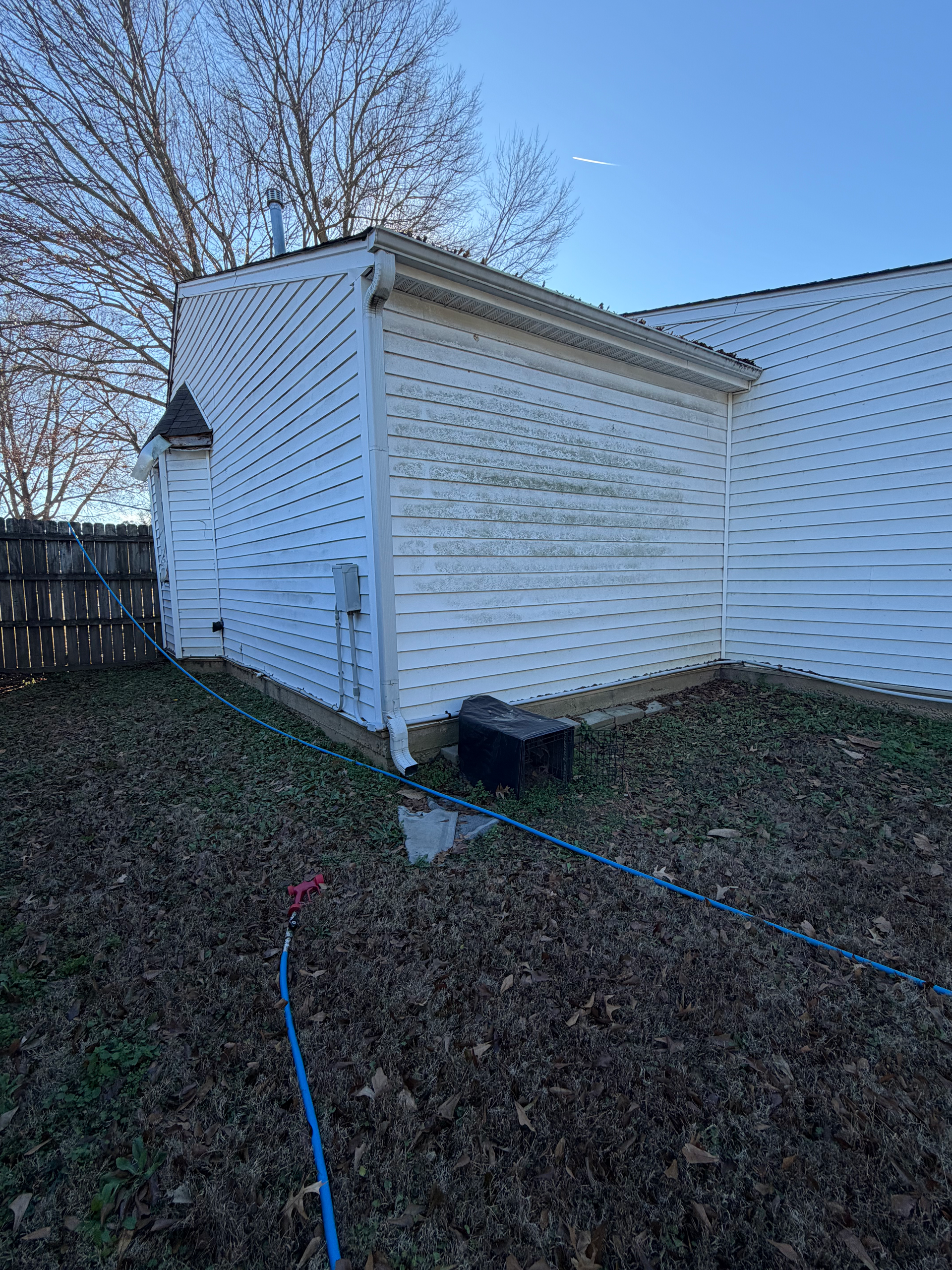 A white-sided exterior wall of a house corner in a backyard filled with dry leaves under a clear blue sky.