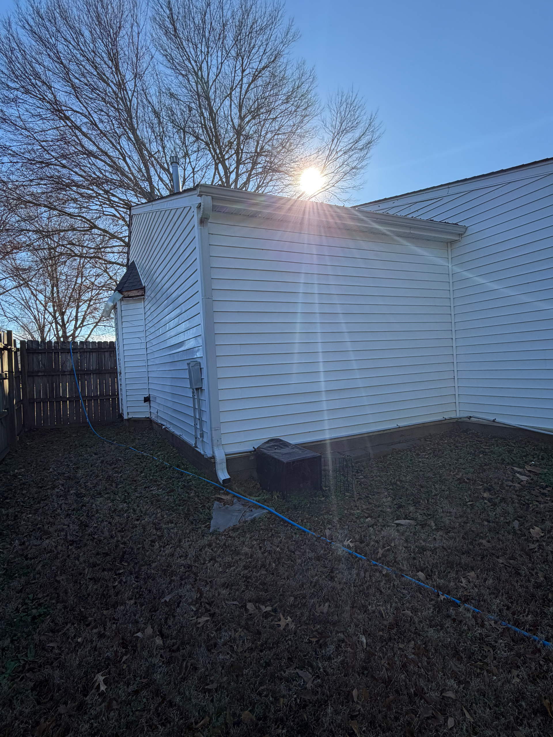 Sunlight shines brightly behind the corner of a white-sided building with a wooden fence and yard in the foreground.