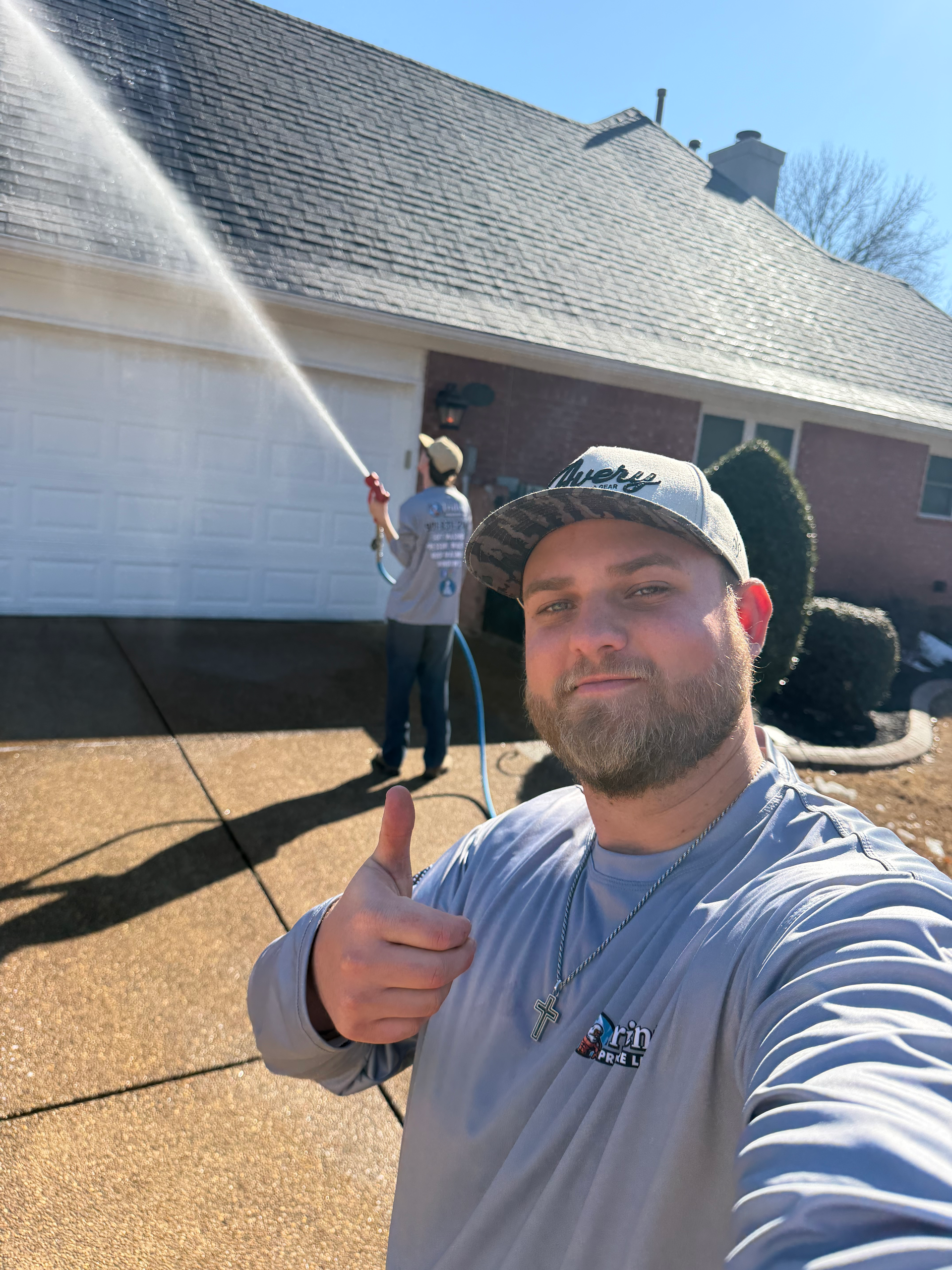 A man wearing a cap and long-sleeved shirt gives a thumbs up while a colleague pressure washes a house roof behind him.