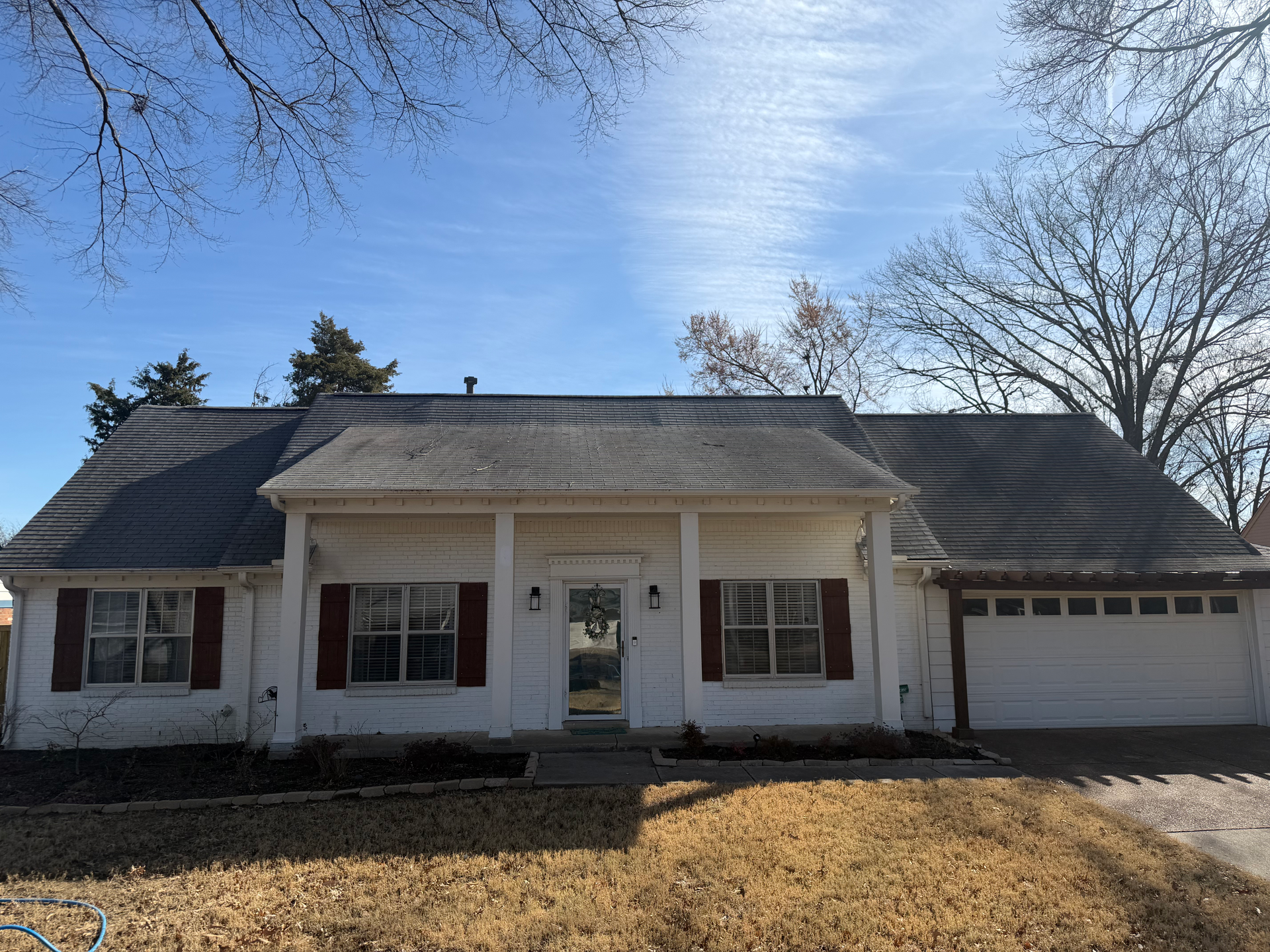 A one-story white house with dark shutters, a covered porch, and an attached garage under a blue sky.