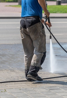 A person in a blue shirt and work pants using a high-pressure washer to clean a paved walkway.