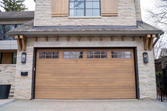 A tan wood-paneled garage door with horizontal slats and rectangular windows, set in a stone facade with wooden brackets.