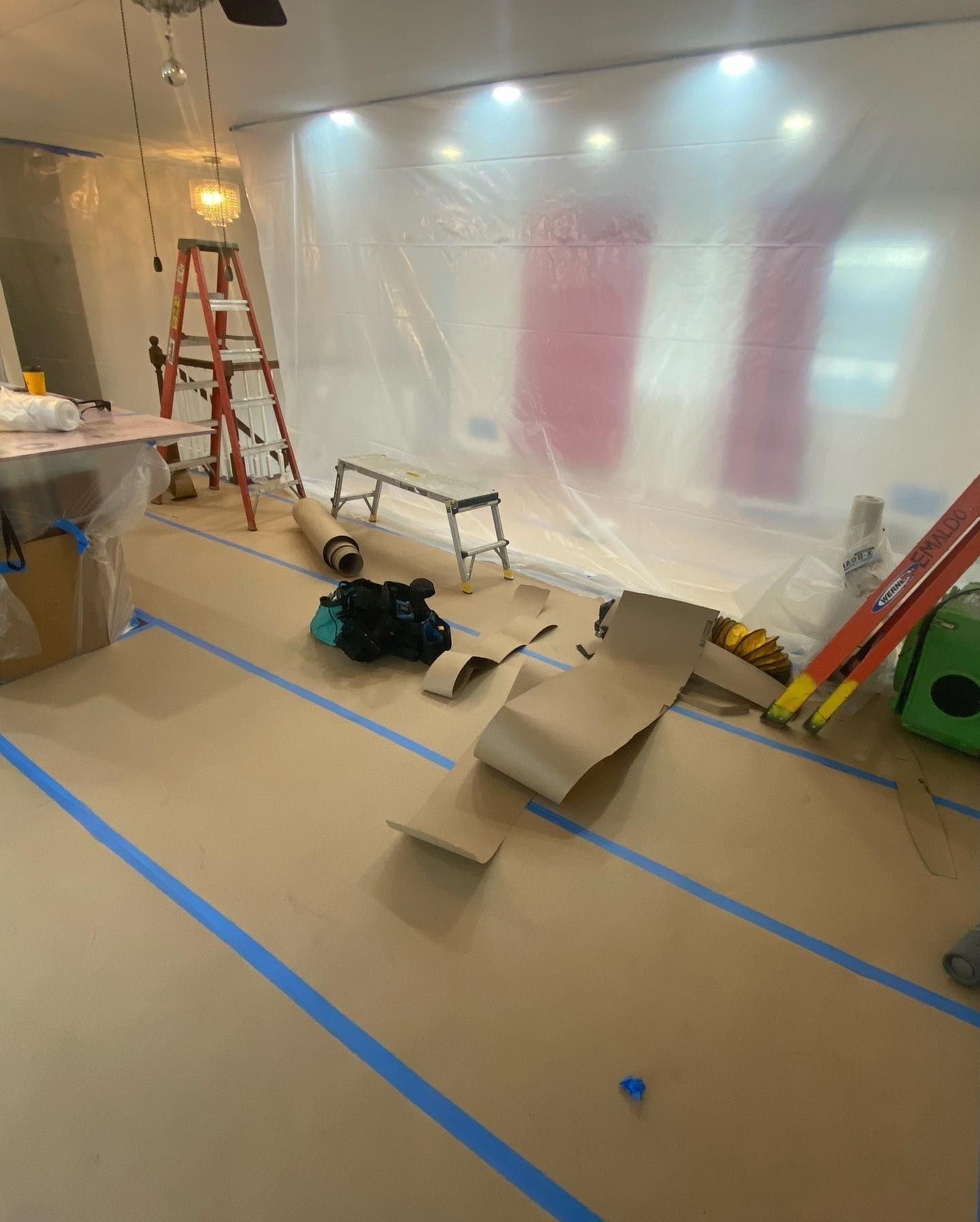 Room under renovation, covered in protective plastic and paper. Ladders, tools, and supplies are scattered about.