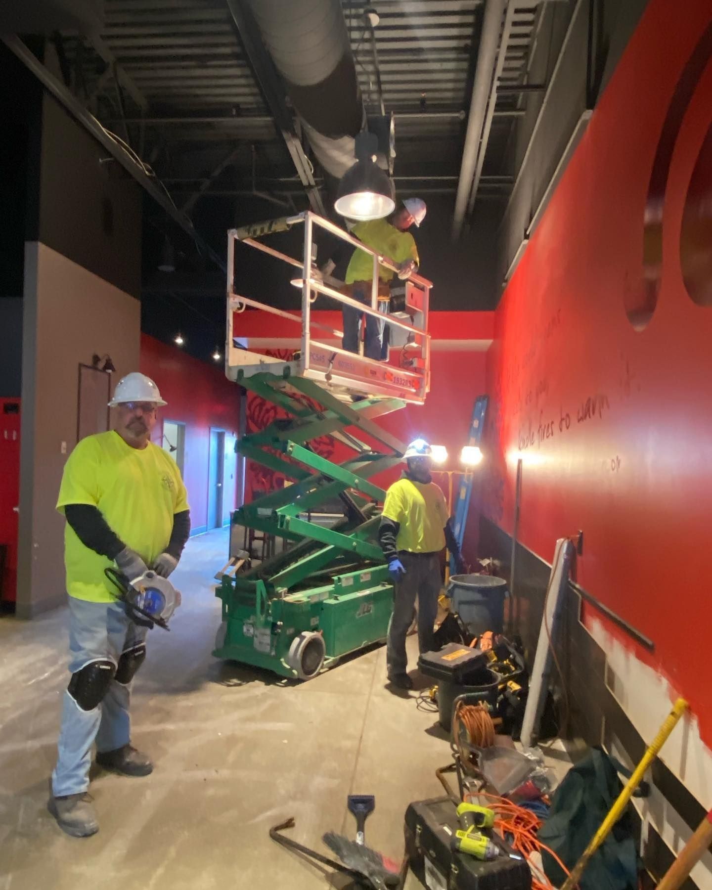 Construction workers using a lift and power tools in a room with red walls and a black ceiling duct.