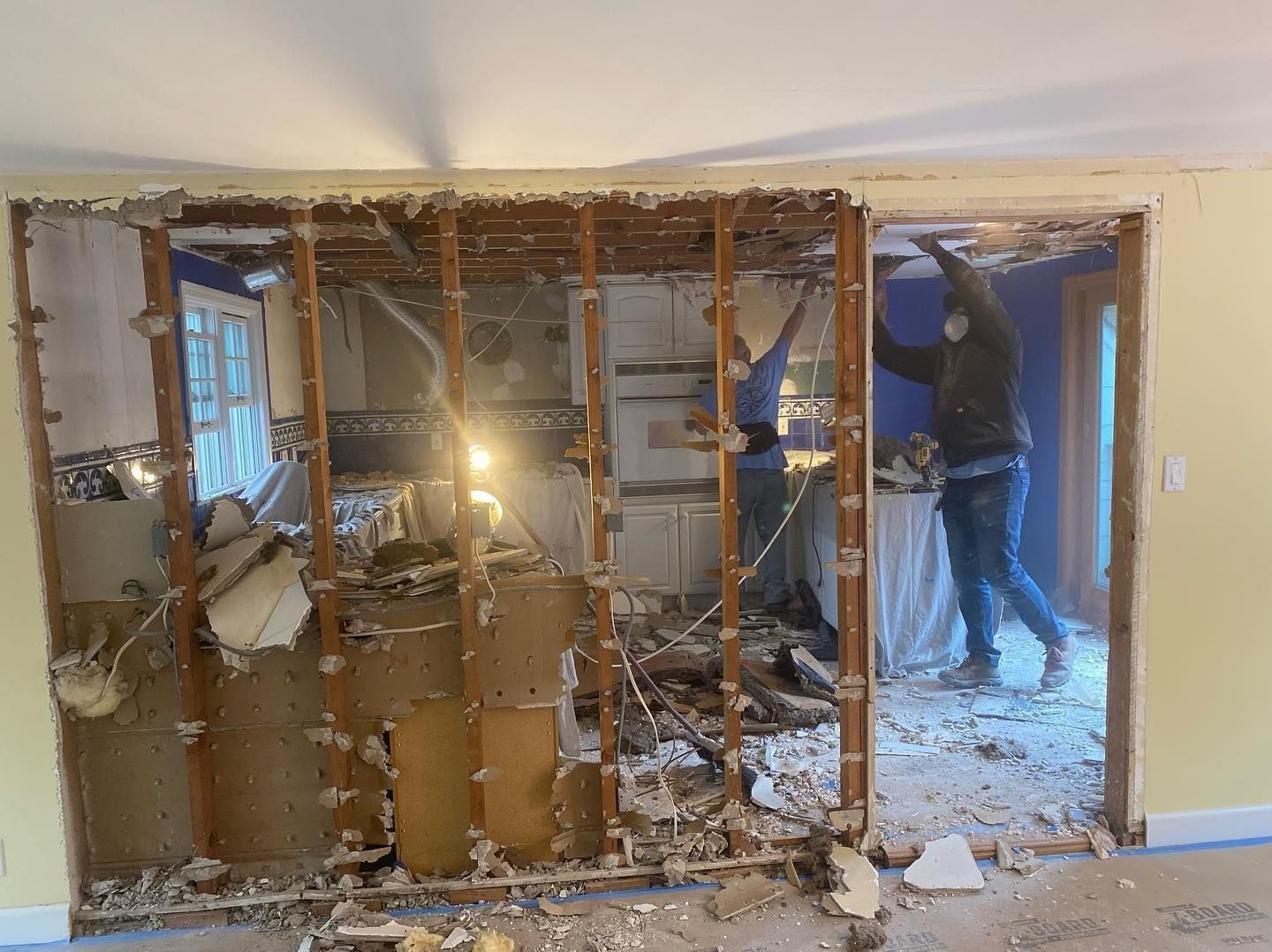 Two people removing a wall in a house, revealing studs. Debris litters the floor.