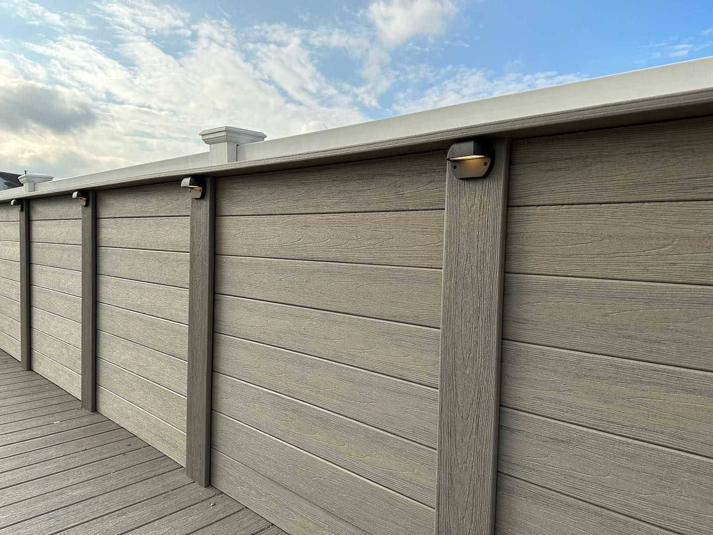 Gray composite fence with vertical posts, deck, and overhead lights against a cloudy sky.