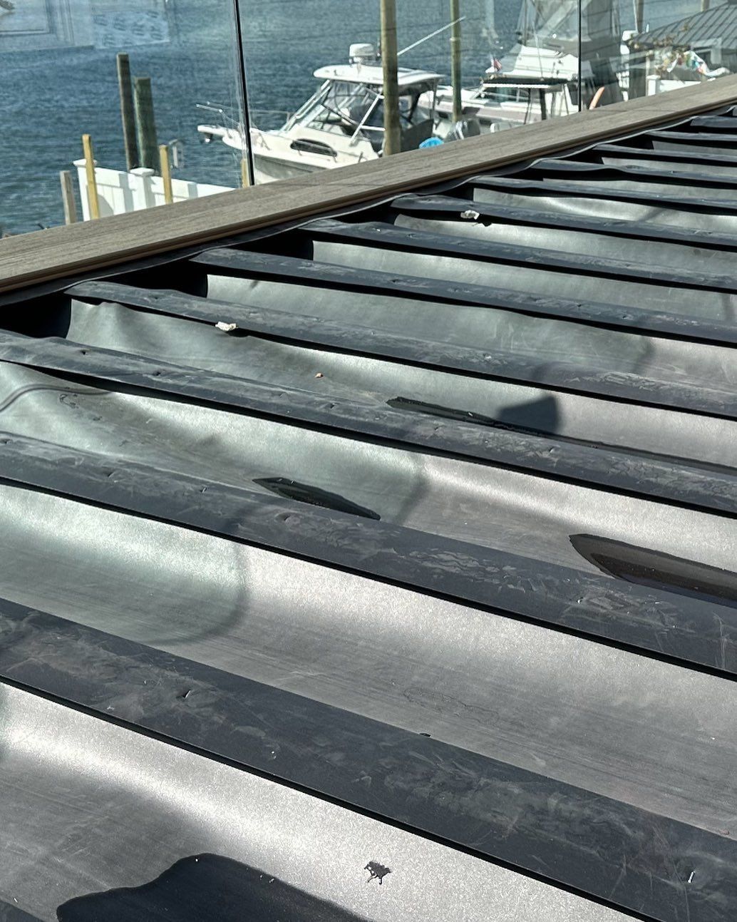 Close-up of a black, corrugated roof with a wooden board on top; boats and water in the background.