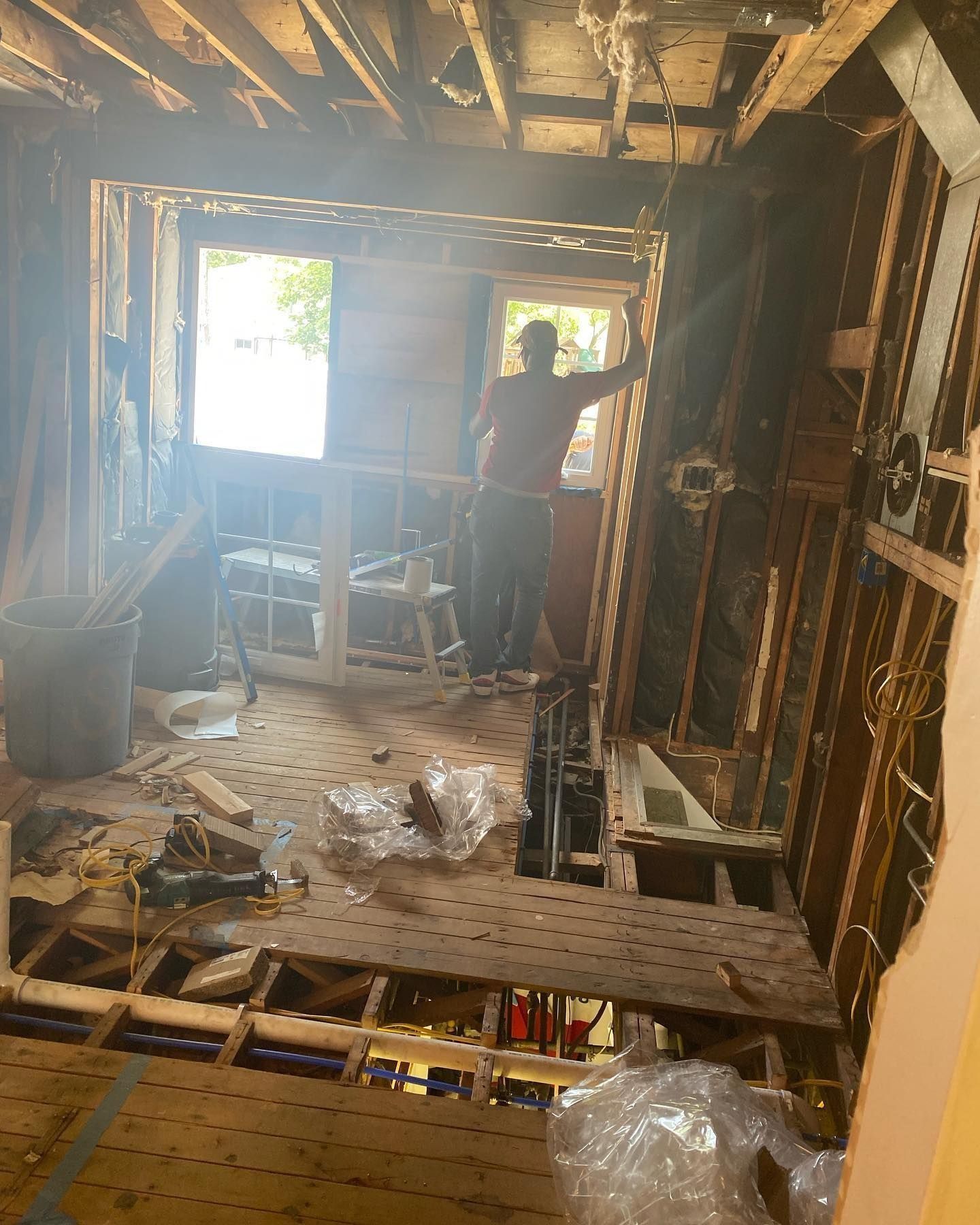Room under renovation; person standing near a window, holding a panel. Visible studs, exposed flooring, and debris.