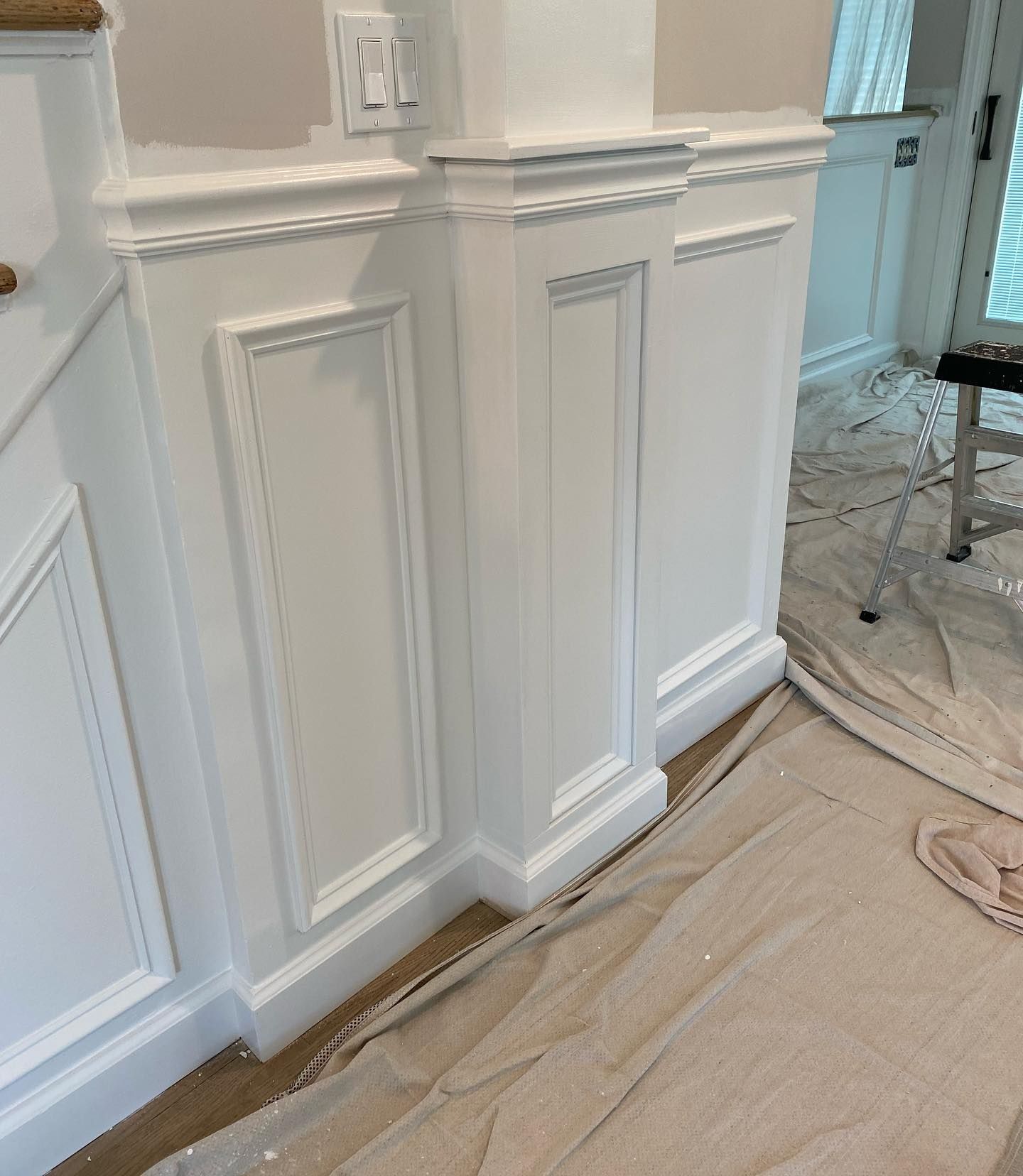 White wainscoting and trim on a wall and pillar with an electrical outlet; a work area with drop cloths.