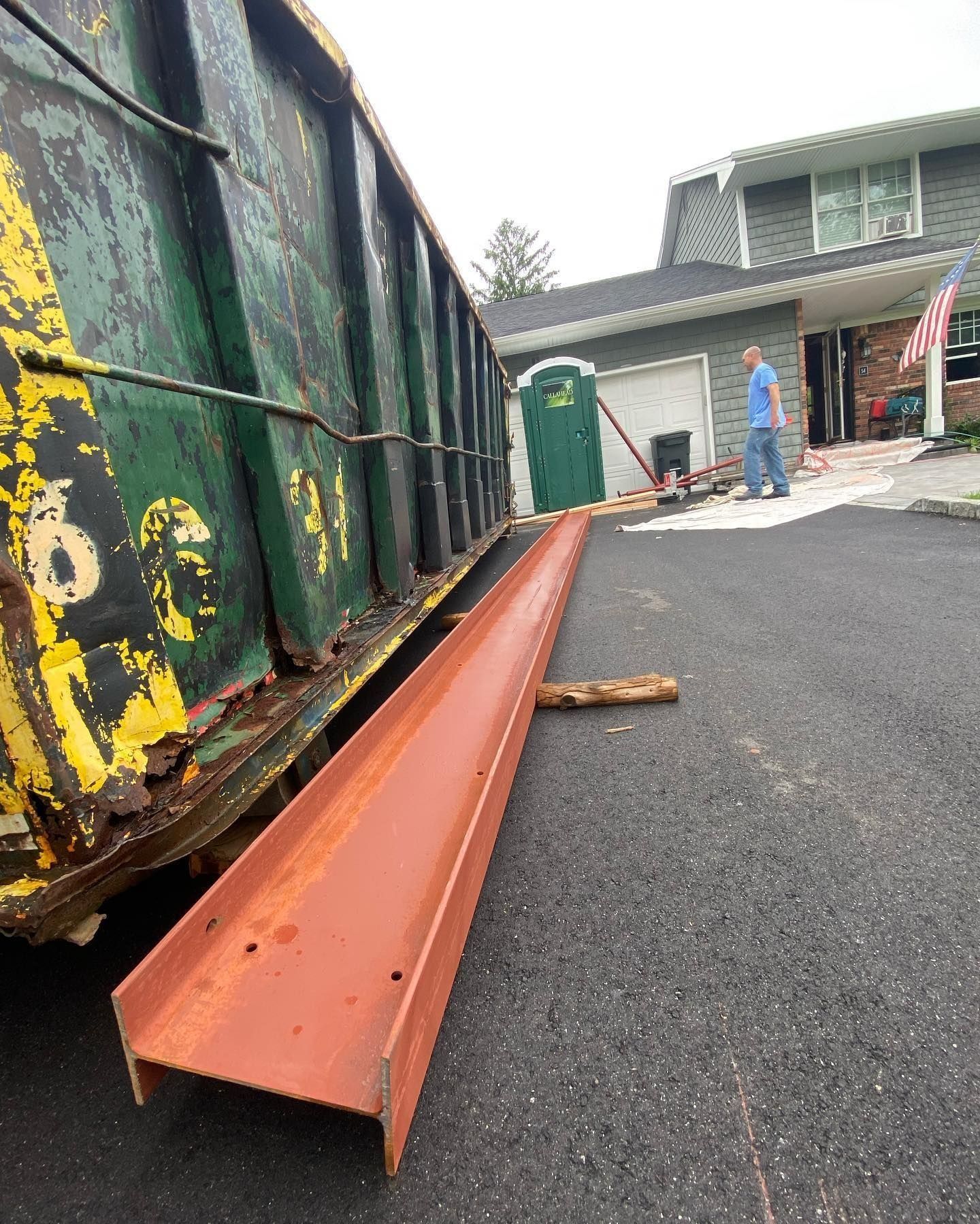 A rusty steel beam acts as a ramp for a dumpster on a residential driveway. A man walks nearby.