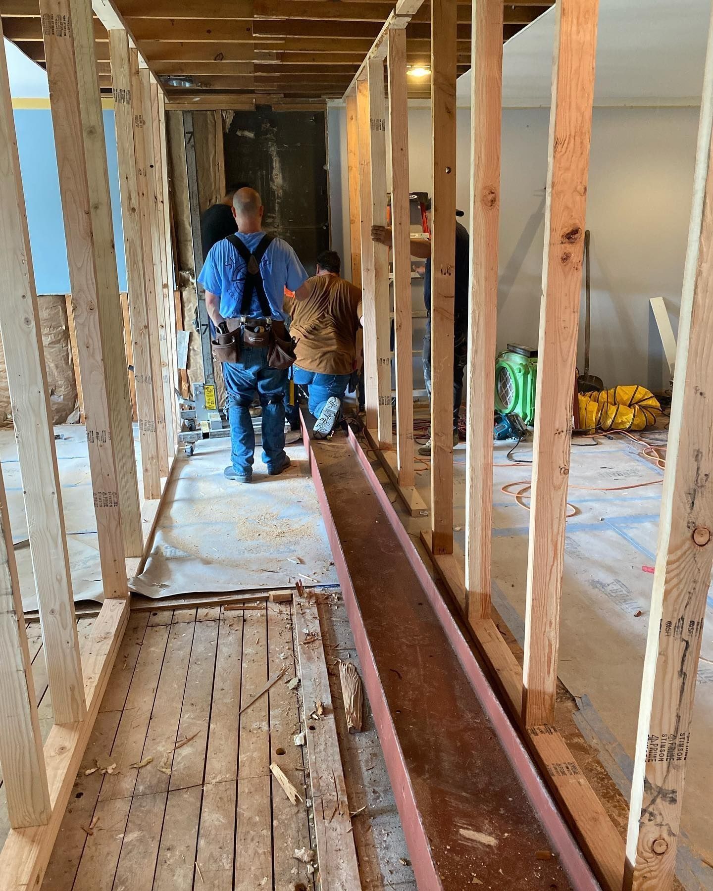 Two construction workers in a hallway with exposed wooden framing and metal beam.