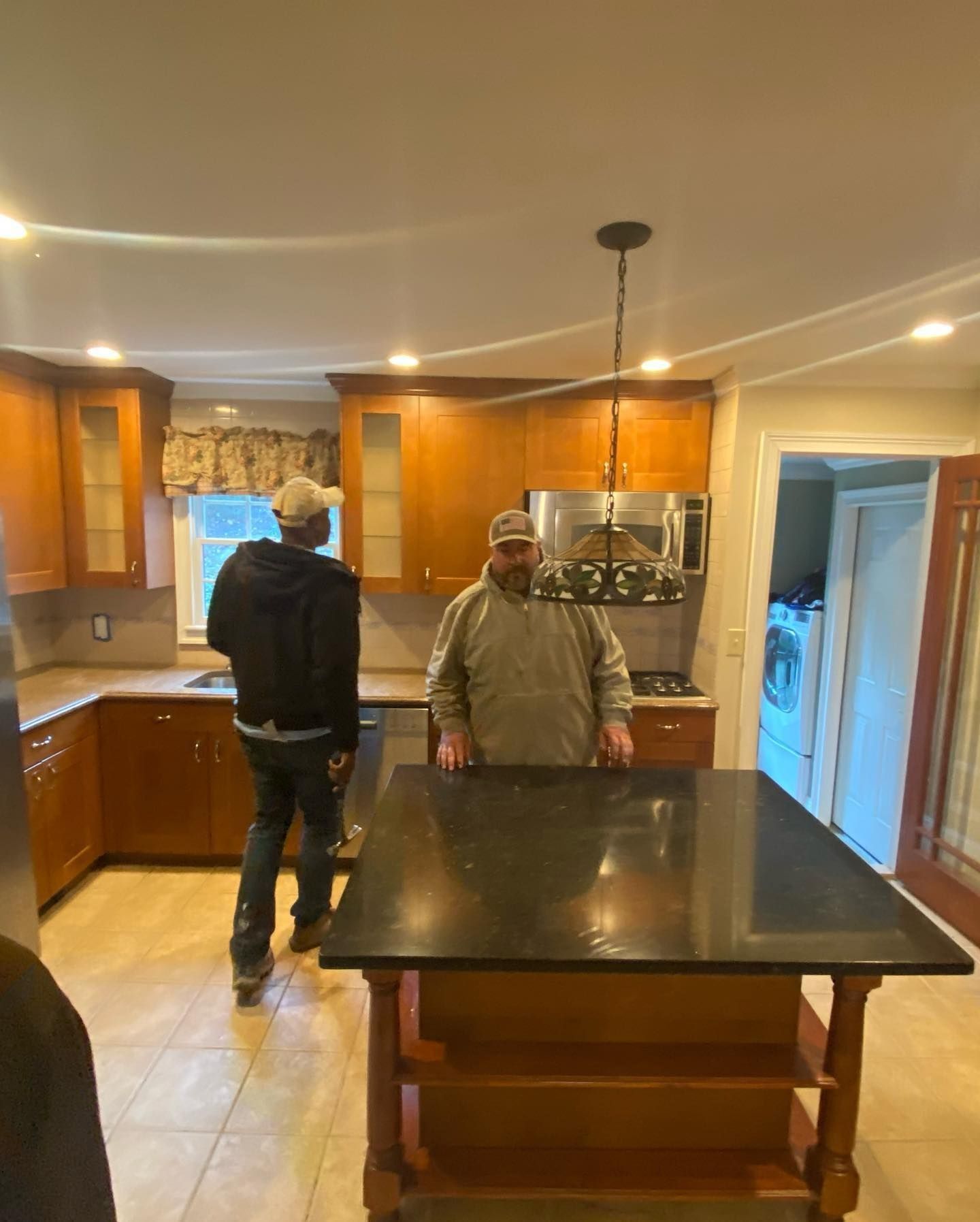 Two people in a kitchen with brown cabinets and a black countertop island. One looks toward a window.
