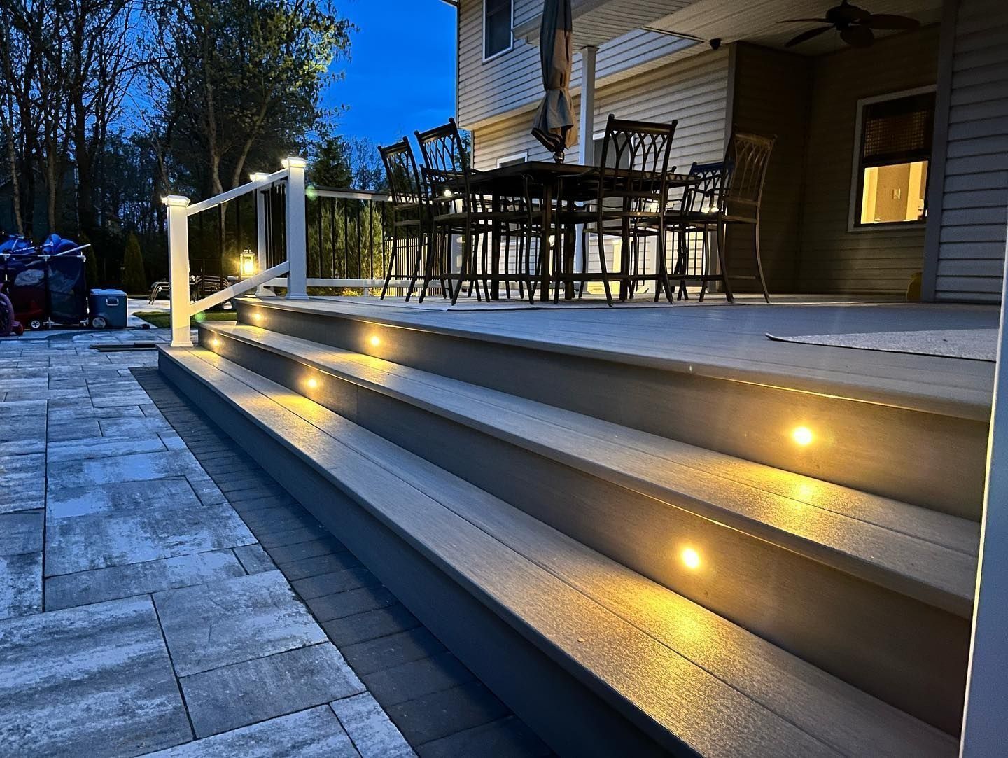 Lit steps lead up to a deck with table and chairs. Deck and railing have lights; dusk setting.