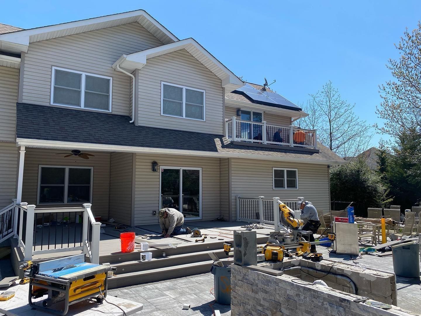 Backyard construction: workers using tools near a house with a deck and balcony on a sunny day.