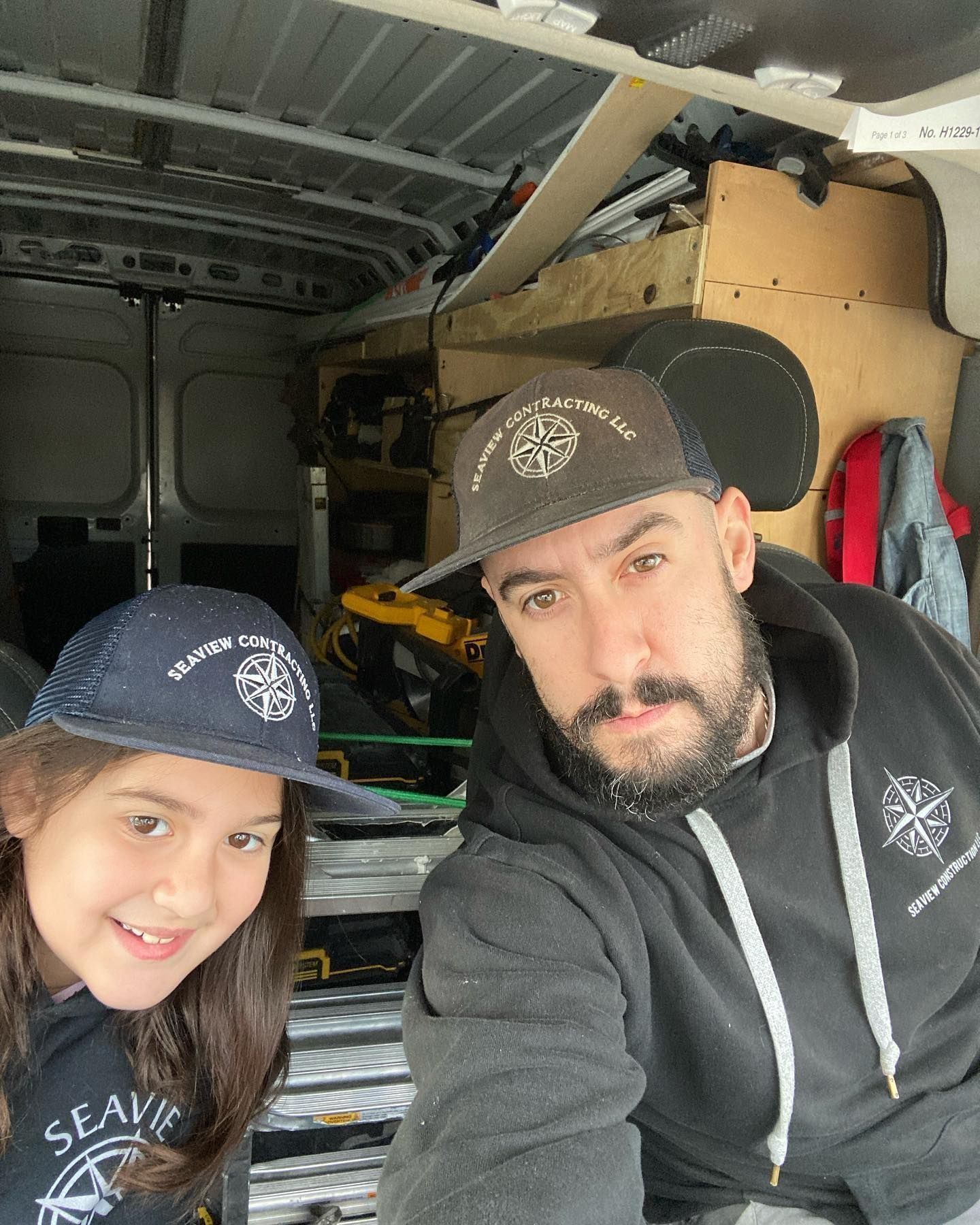 Man and girl in matching hats inside a van, smiling. Grey hoodie and cap, dark interior.