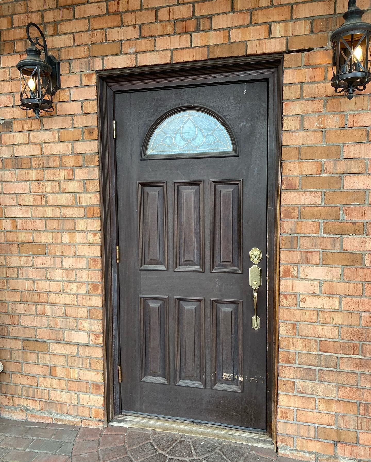 Brown door with arched glass panel, set in brick wall with two outdoor lights.