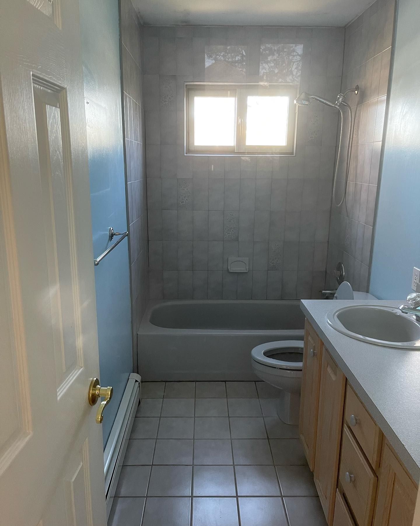 Bathroom with light blue walls, tiled floor, white tub, toilet, vanity, and window.