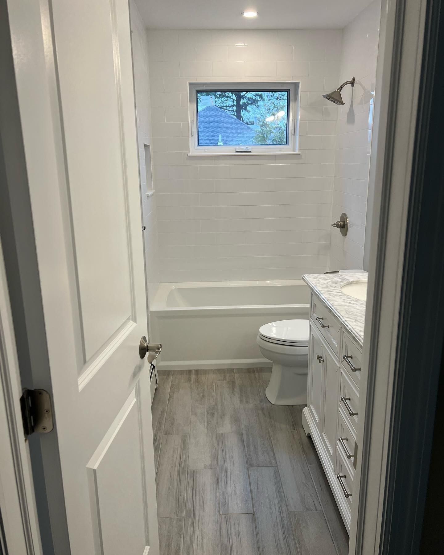 Bathroom with white tile walls, wood-look floor, bathtub, toilet, vanity, and small window.