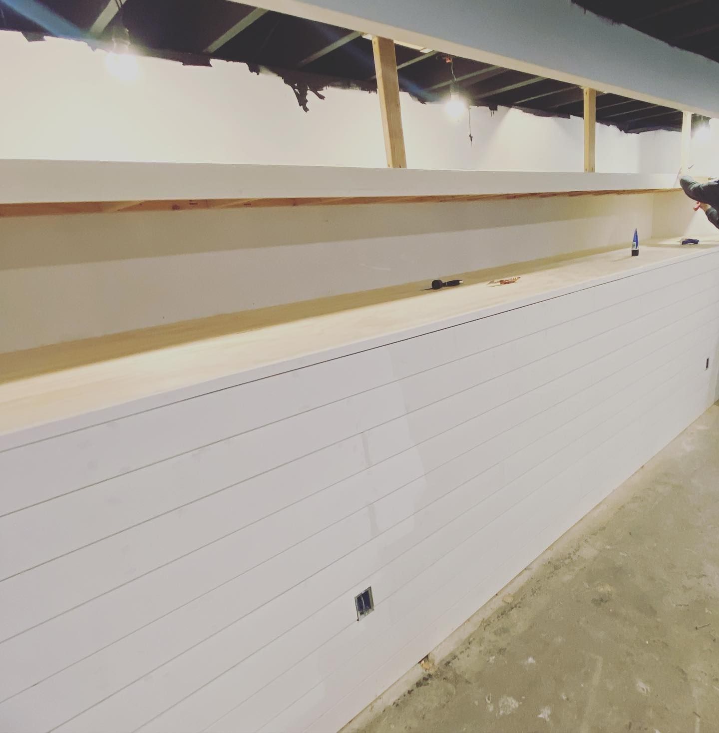 White-paneled wall with a long wooden shelf above it, under a dark ceiling during construction.