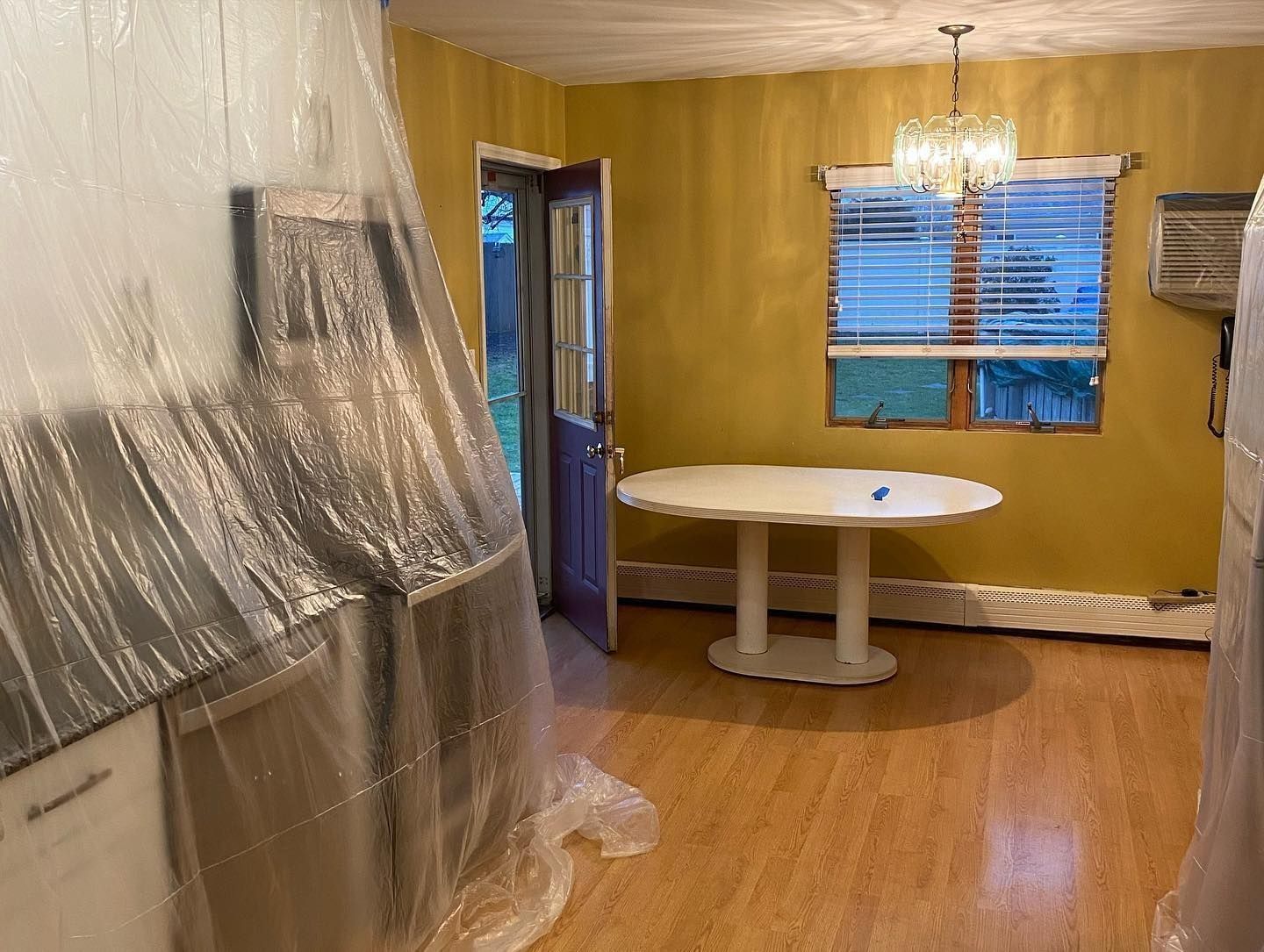 Dining room: white table, gold walls, window with blinds, plastic sheeting covering appliances.