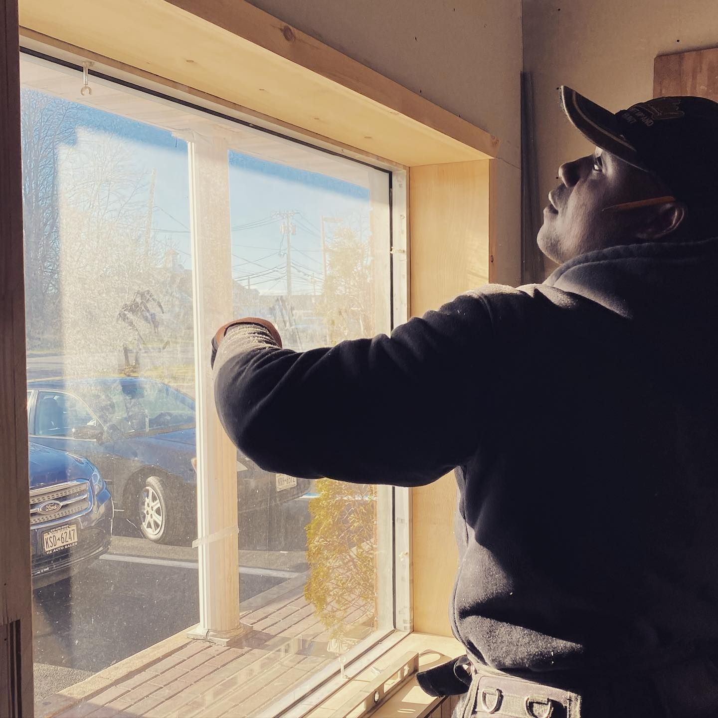 Person installs window trim. Indoors, natural light streams in. They wear a cap, hoodie, and gloves.