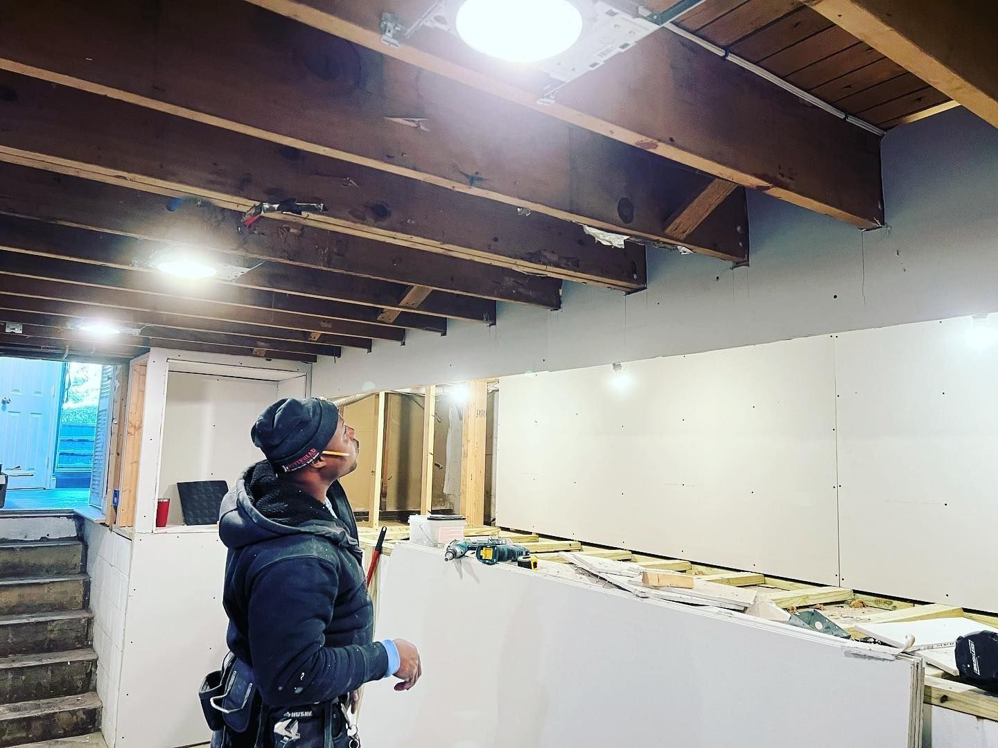 A person in a basement surveys a wall during renovation, with exposed beams and drywall.