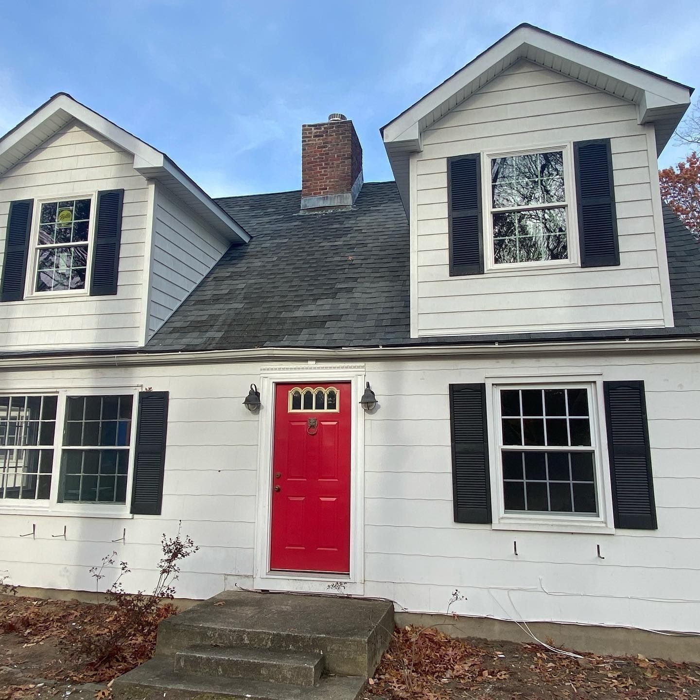 White house with red door, black shutters, two dormers, and a chimney.
