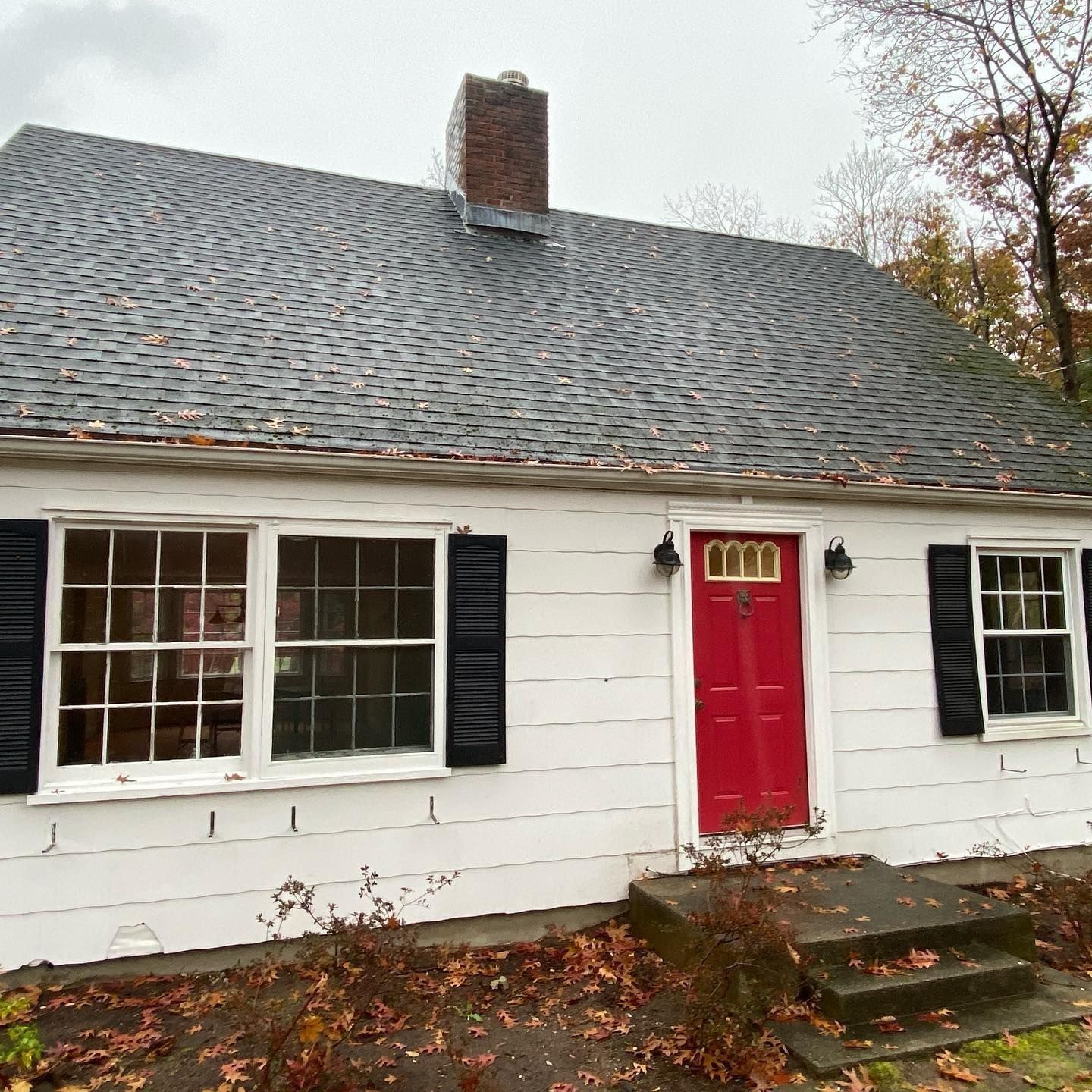 White house with black shutters, red door, and dark gray roof. Chimney on roof.