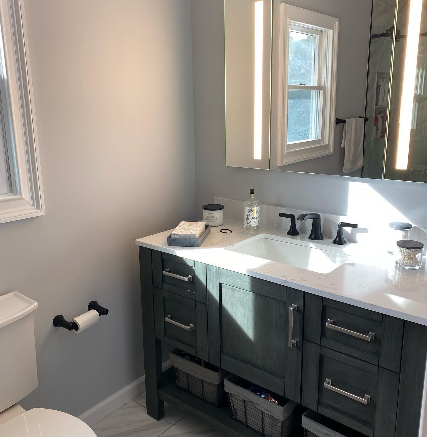 Bathroom with gray walls, dark vanity, white sink, and window. Sunlight highlights the counter.
