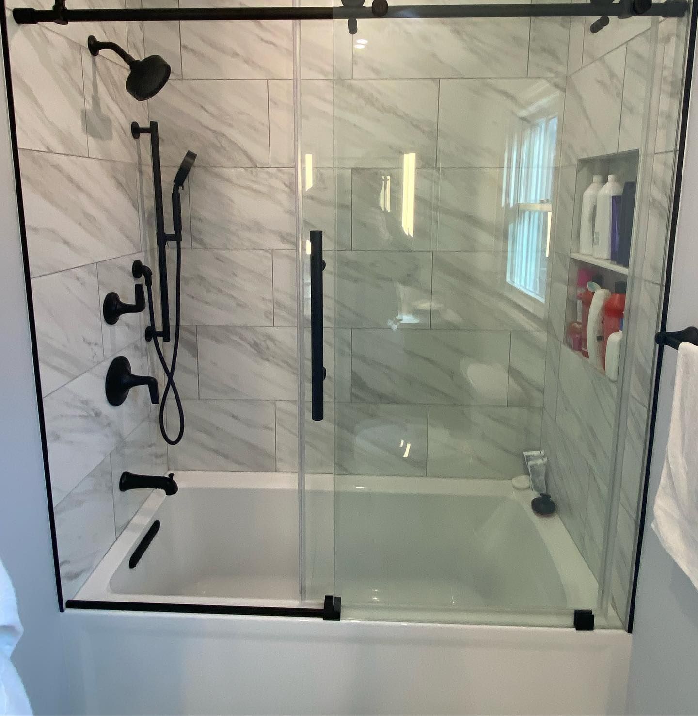 Black framed glass shower enclosure over a white bathtub with marble tile walls and black fixtures.