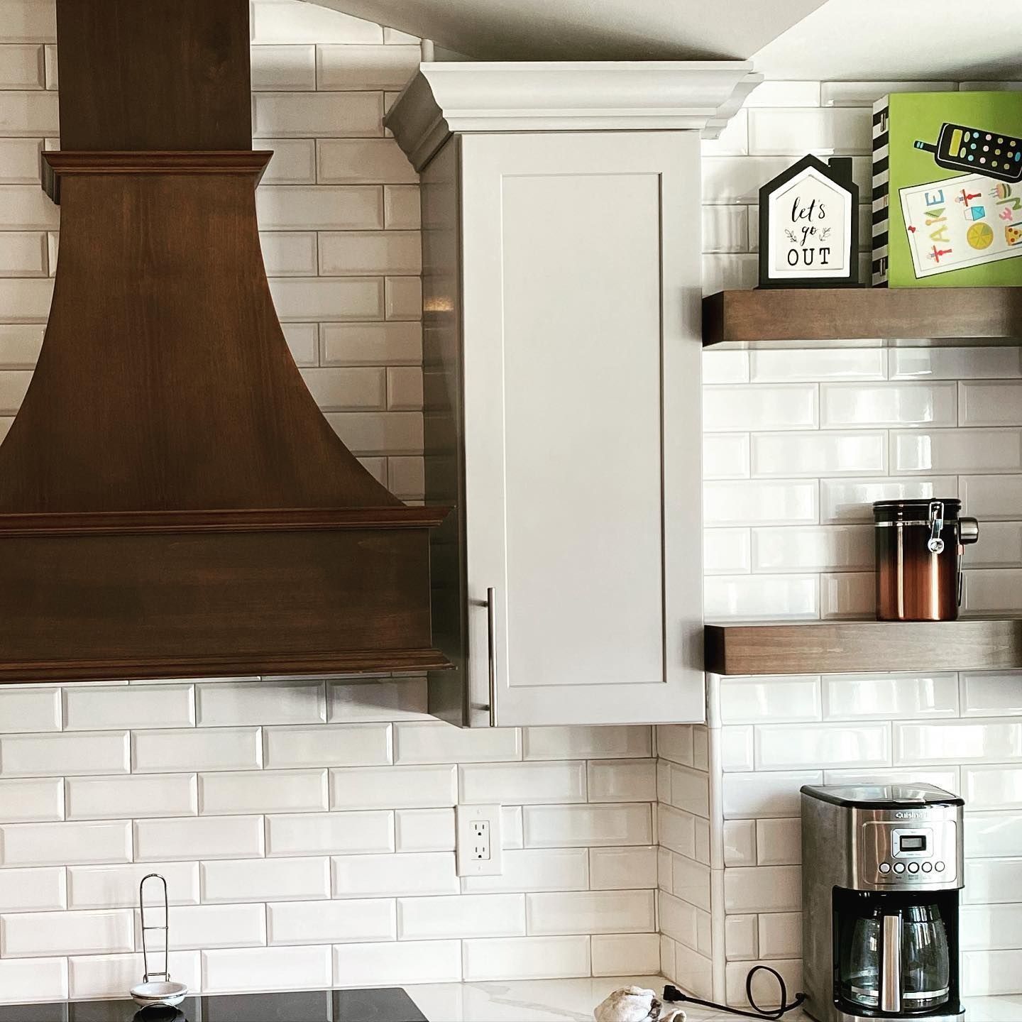 Kitchen with white tile backsplash, wooden hood, white cabinet, floating shelves, and coffeemaker.