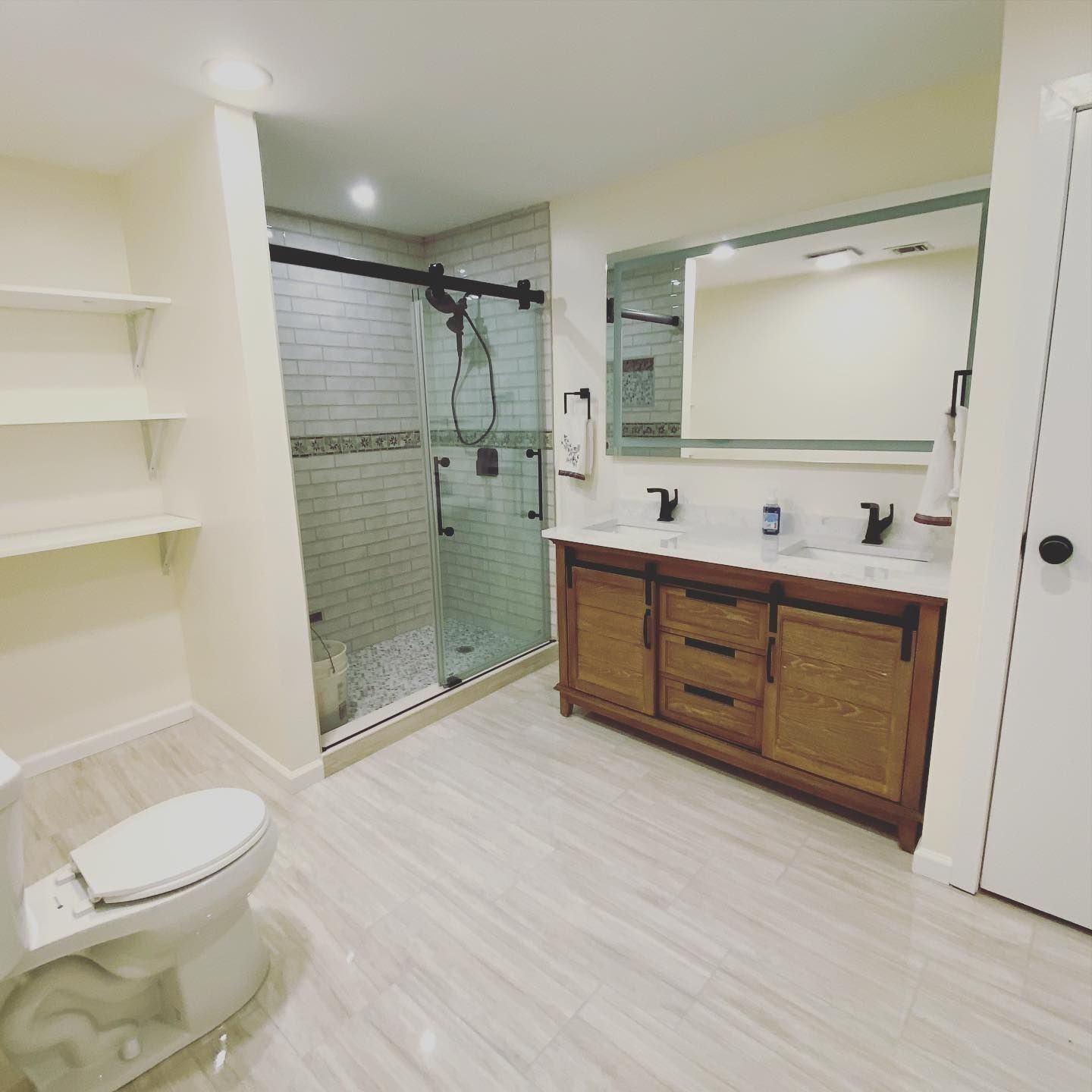 Bathroom with glass shower, double vanity, toilet, and shelving. White walls and light wood accents.
