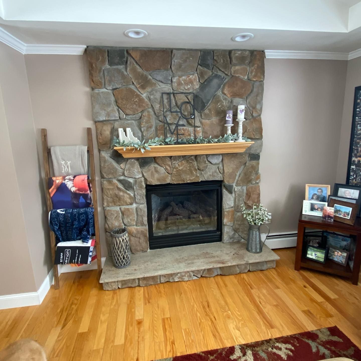 Stone fireplace with wooden mantel, flanked by a leaning blanket ladder and small side table with photos.