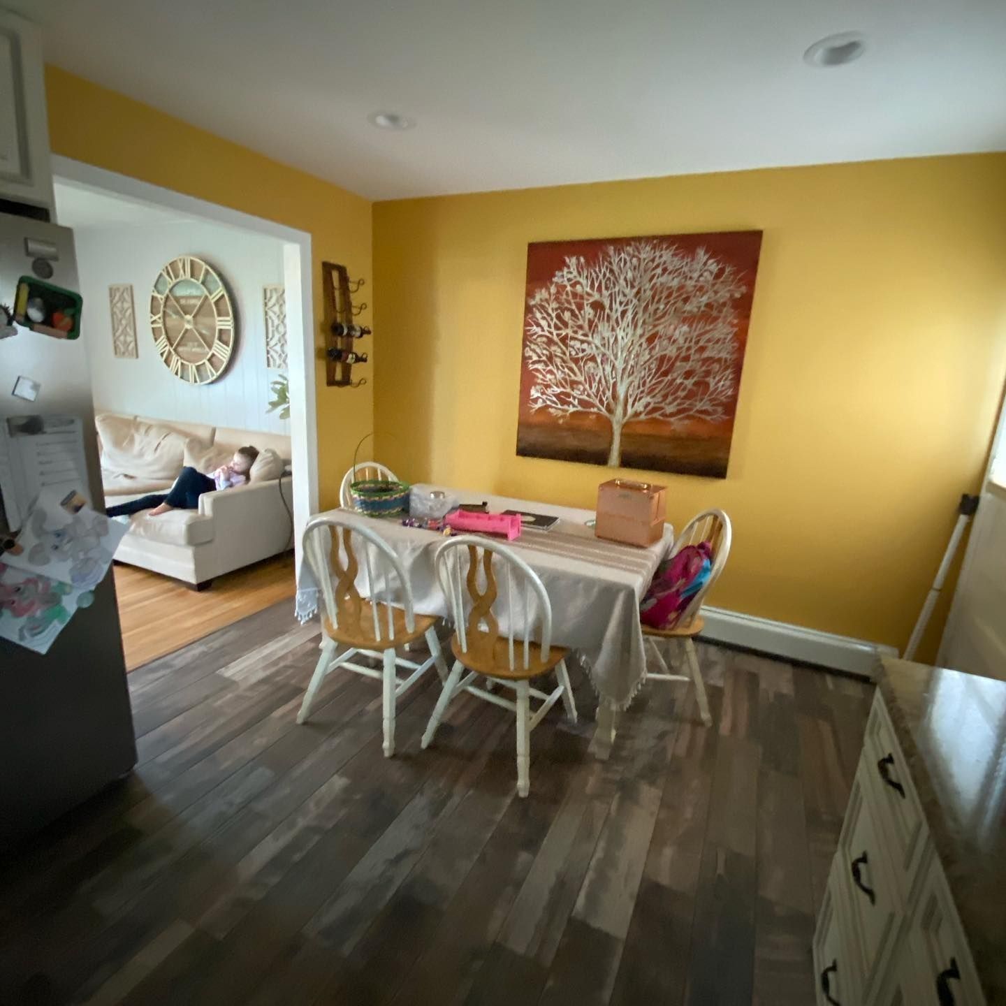 Dining room with table, chairs, art, and doorway to living room with person on couch; yellow walls, wood floor.