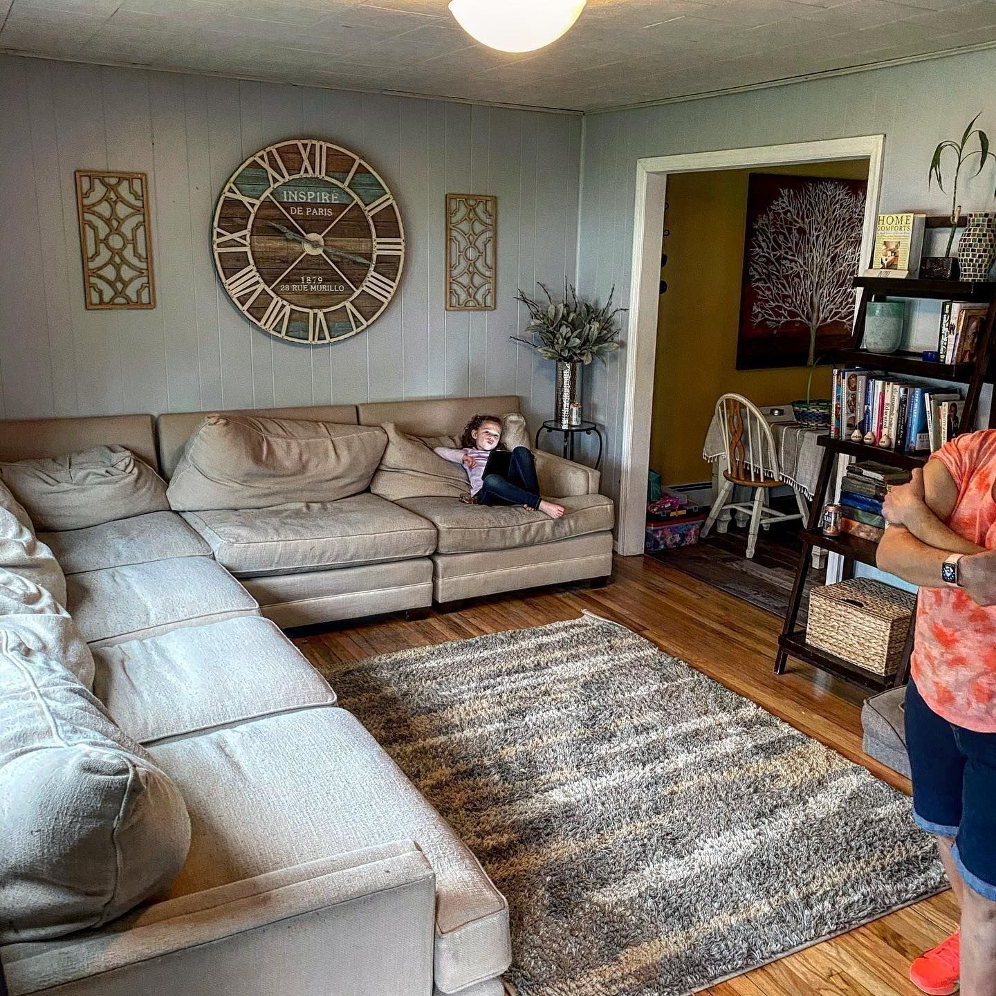 Living room with sectional sofa, rug, clock, person sitting on sofa, bookshelves, and person standing.