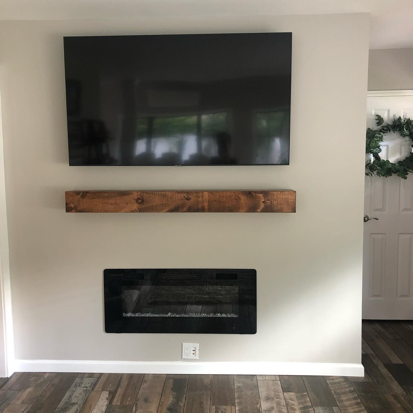Wall-mounted TV, wooden shelf, and fireplace; white door with wreath and wood flooring.