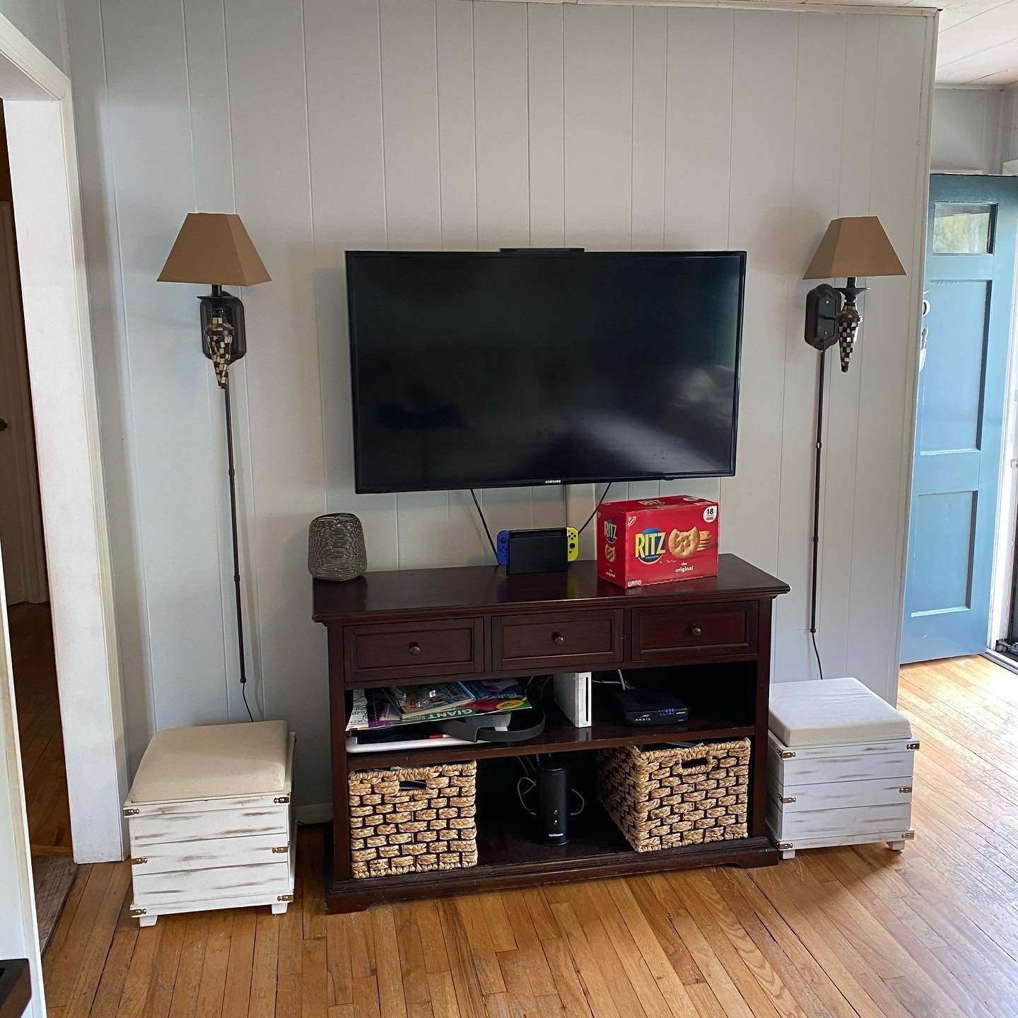 TV mounted above a dark brown cabinet with baskets, surrounded by wall lamps and ottomans, against a white panel wall.