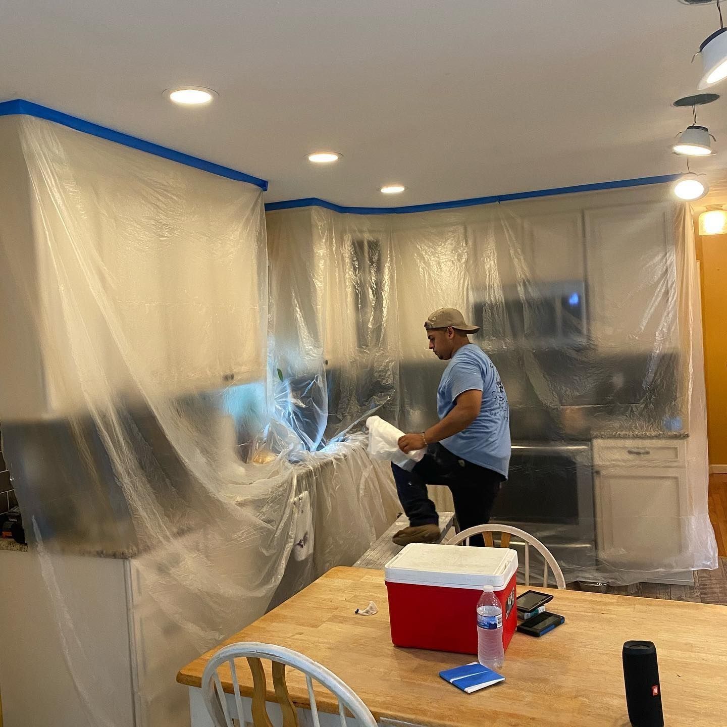 Person painting kitchen cabinets, covered in plastic, while standing on a small ladder.