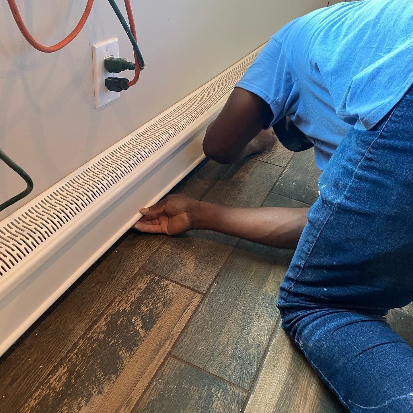 Person kneeling, installing baseboard heater against a white wall. Electrical cords and outlets visible. Wooden floor.