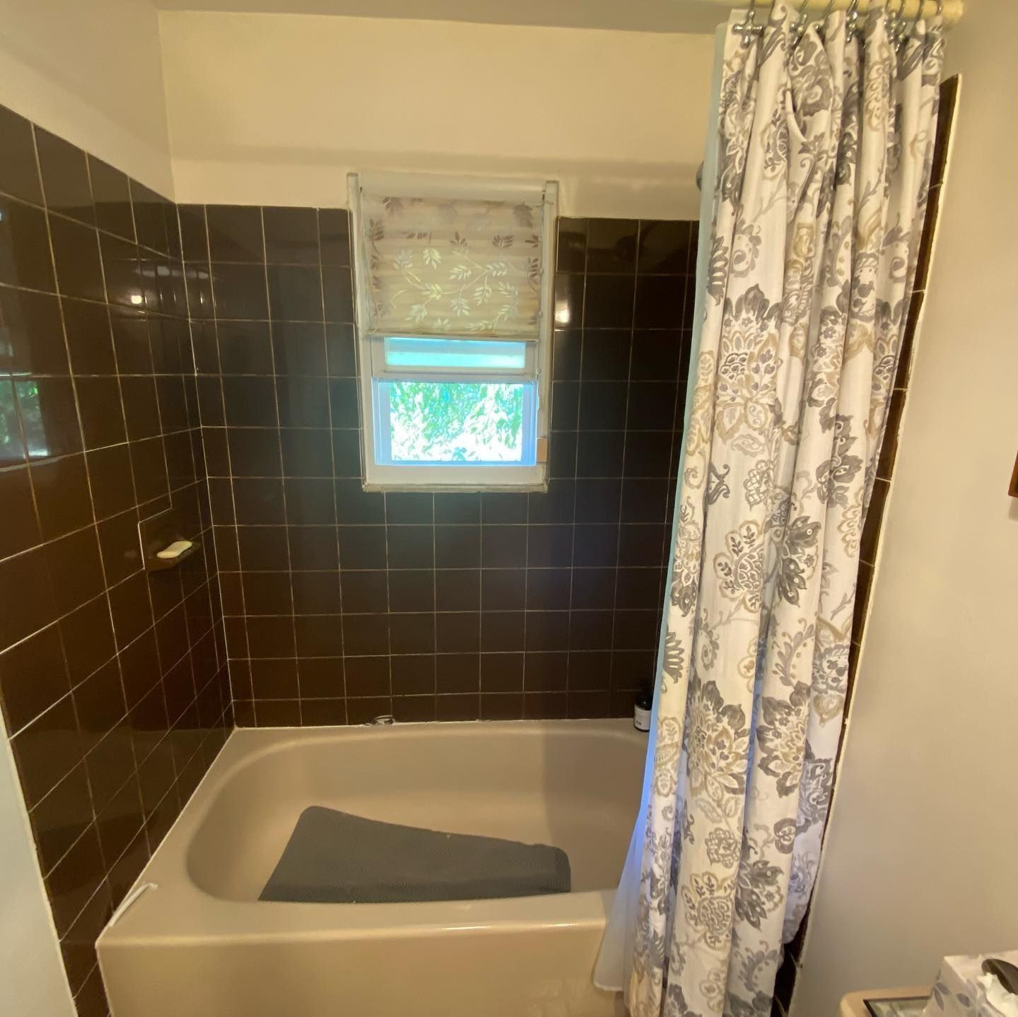 Brown-tiled bathroom with a beige tub, patterned shower curtain, and a small window with a shade.