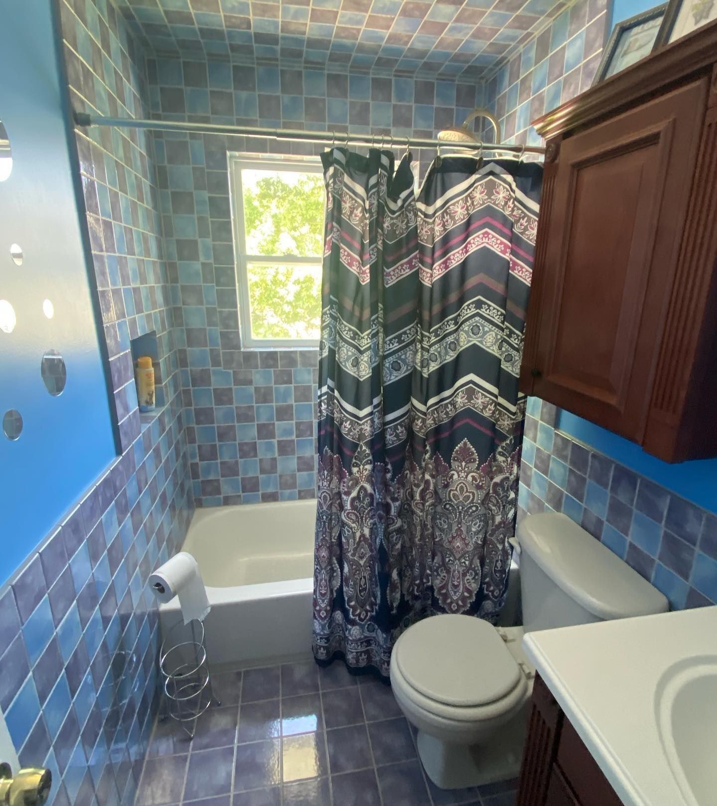Bathroom with blue tiled walls and floor, white tub, toilet, and patterned shower curtain.