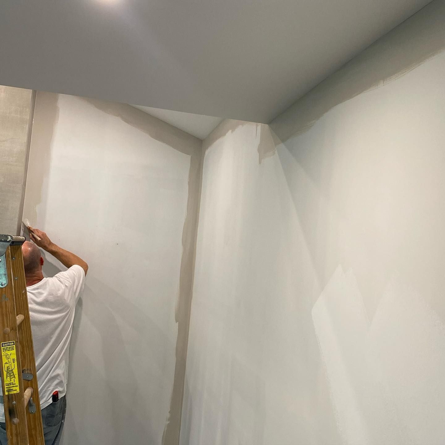 Person on a ladder painting a corner of a room, walls are white, trim is gray.
