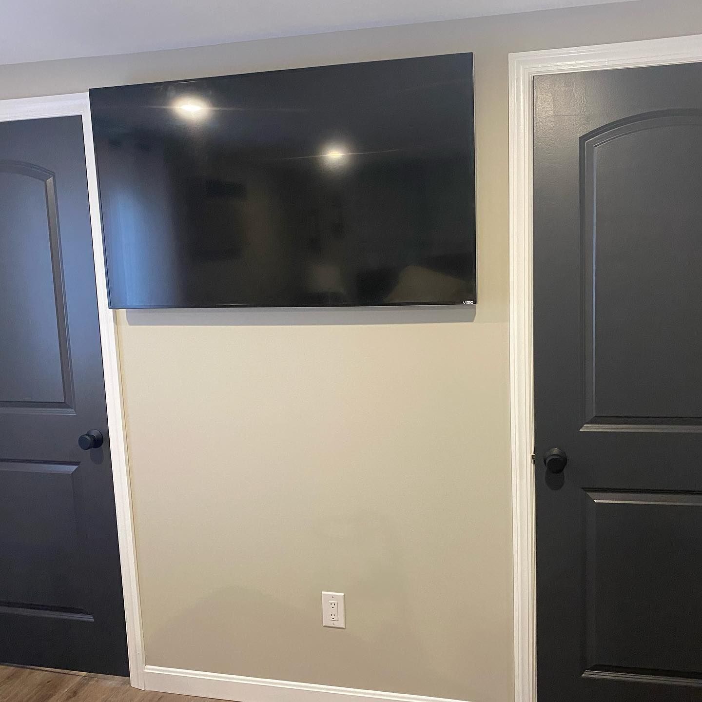A large TV mounted above an electrical outlet on a beige wall, flanked by two black doors.